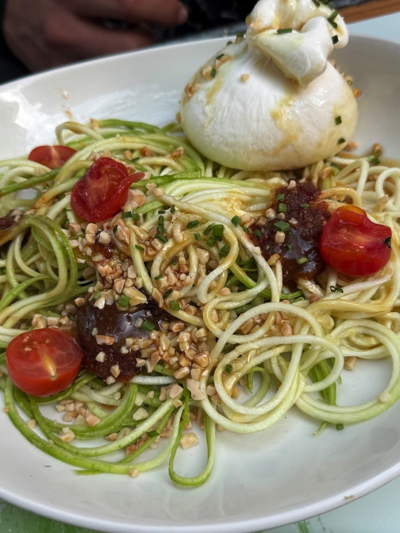 Bowl of spiralized zucchini noodles topped with cherry tomatoes, crushed nuts, dollops of sauce, and a whole burrata, offering a vibrant and refreshing presentation of a modern Mediterranean dish.