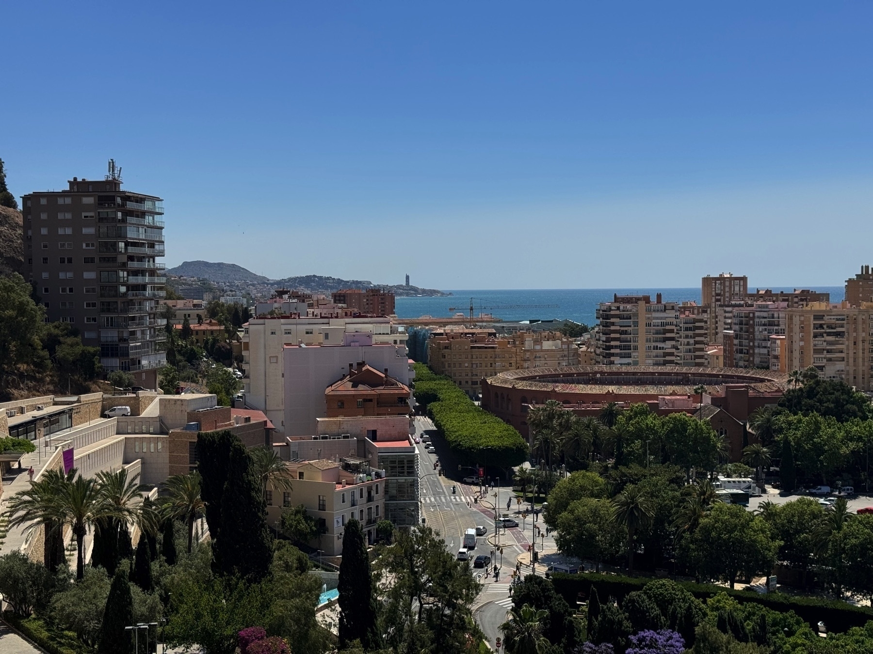 A cityscape view from above looking toward the Mediterranean Sea, with a circular bullring, tree-lined streets, and apartment buildings in the foreground. Hills and a distant lighthouse are visible along the horizon.