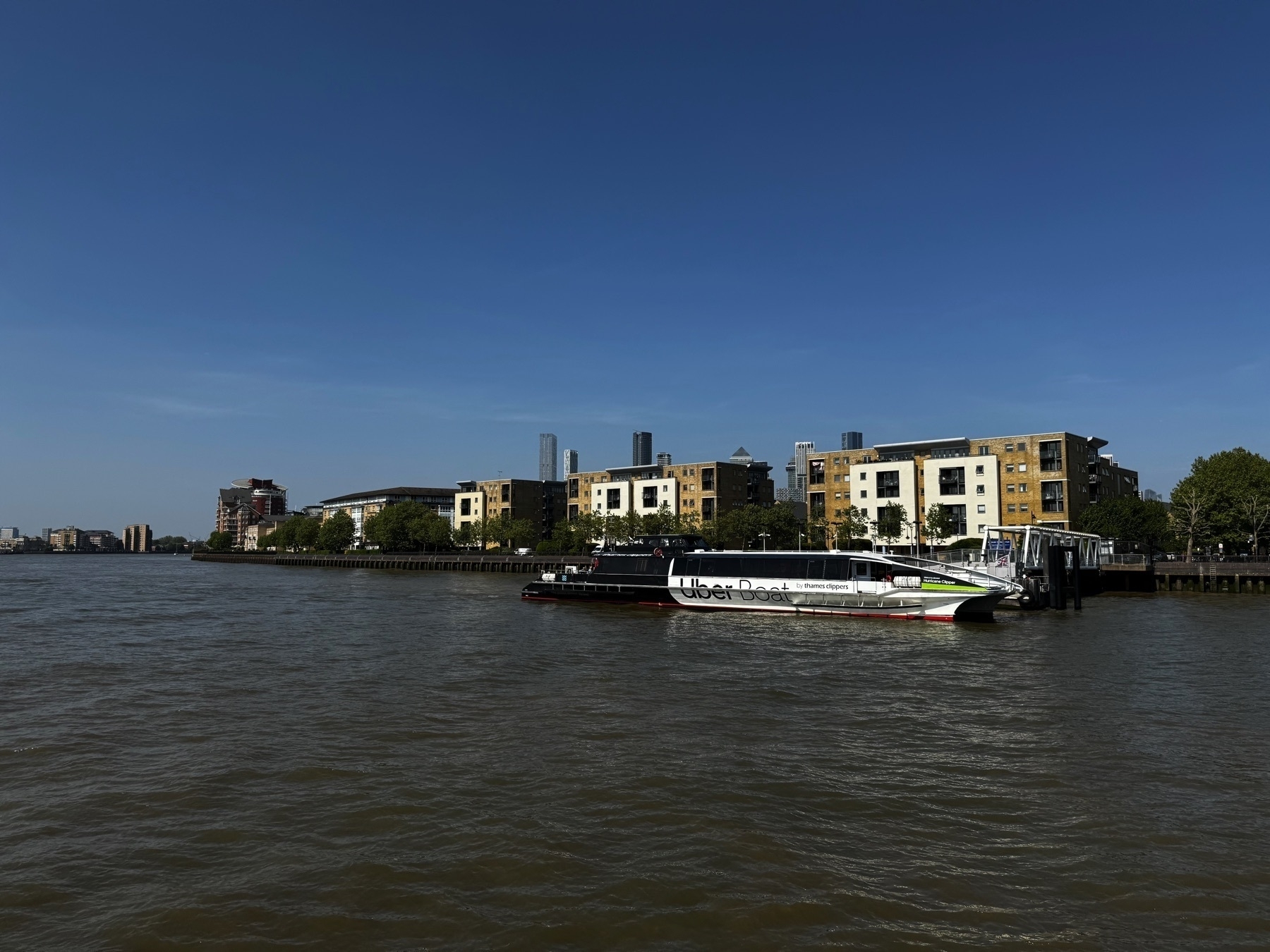 A moored Uber Boat by Thames Clippers sits at a dock along the Thames. Behind it, mid-rise residential buildings with light-colored façades line the riverbank under a clear blue sky.