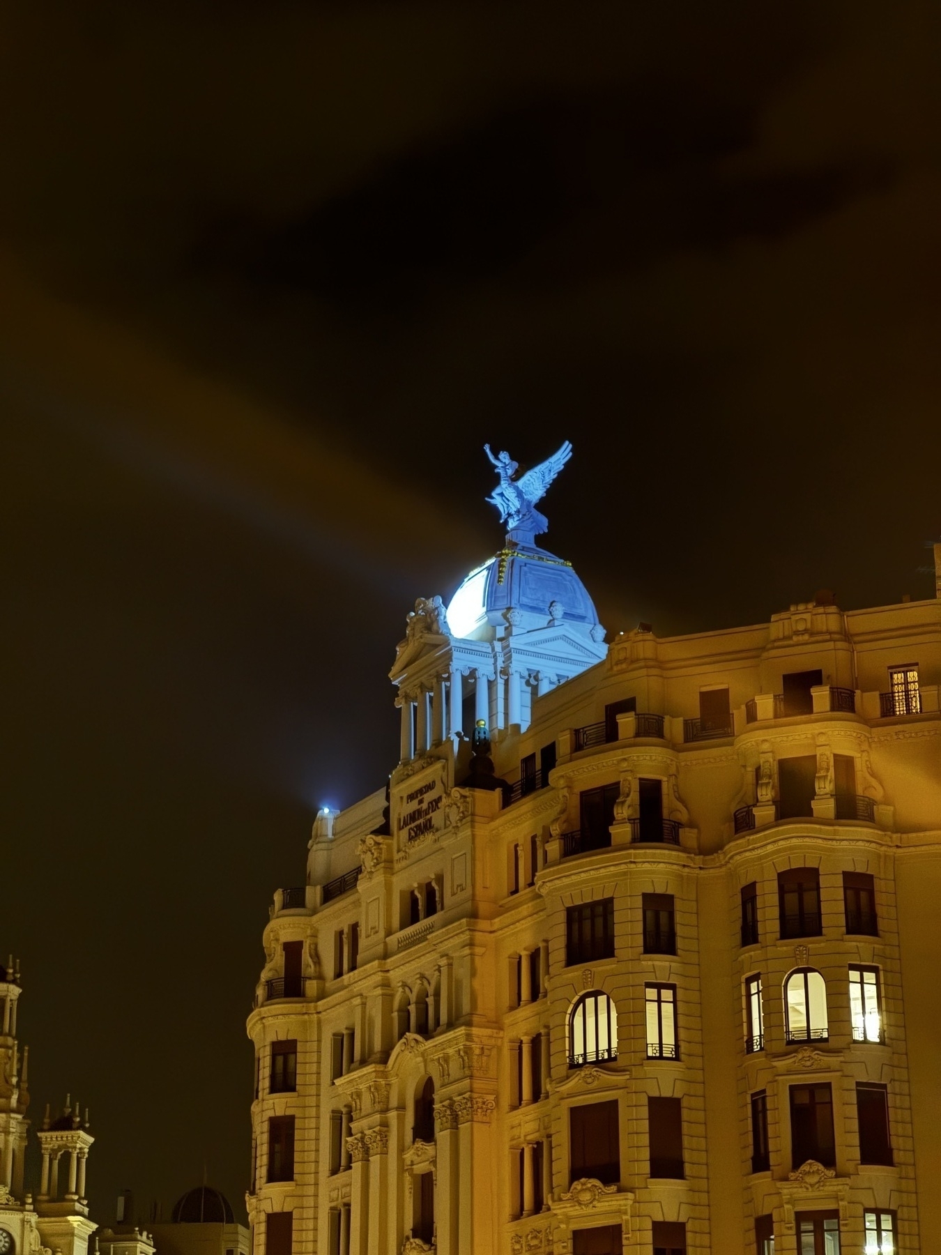 The illuminated dome of the Metropolis Building, topped with a winged statue, shines brightly against the night sky. Its dramatic lighting and classical detailing evoke a sense of grandeur and urban romance.