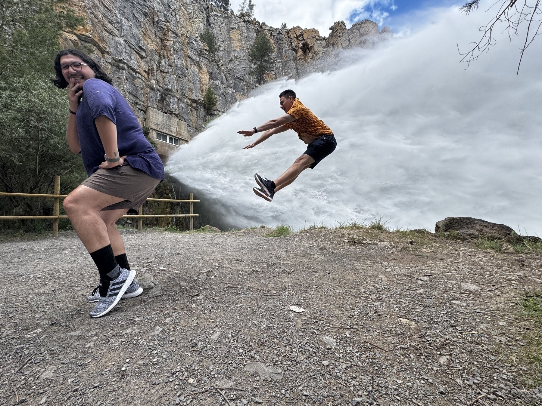 Two people posing playfully in front of a massive water release from a dam; one stands in a cheeky pose while the other leaps mid-air as if blasted by the water jet coming out of the first person’s butt.