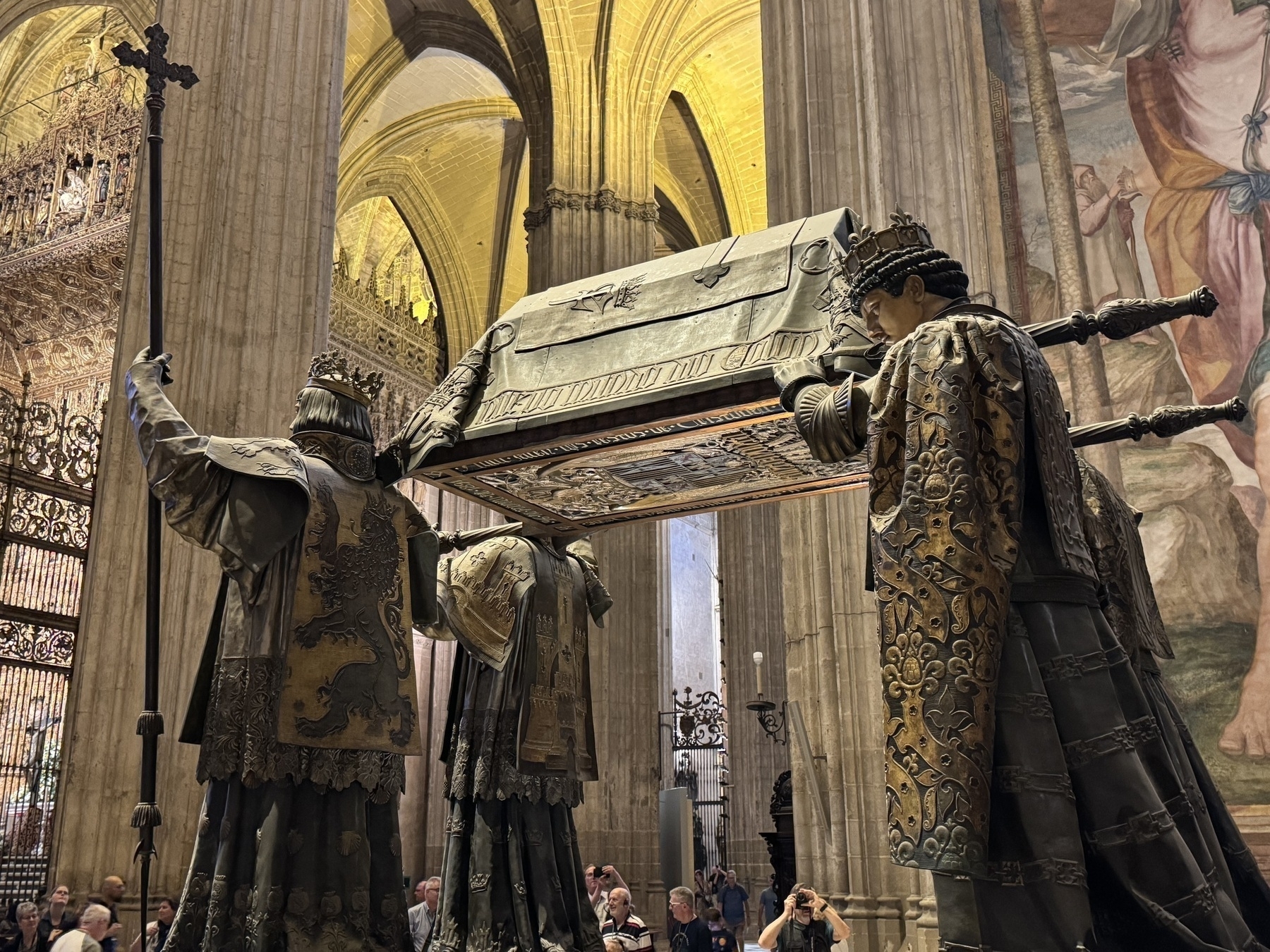 Dramatic sculpture of four heraldic figures bearing the ornate tomb of Christopher Columbus, set within the cathedral’s vast interior under vaulted ceilings, with visitors gathered around the base.