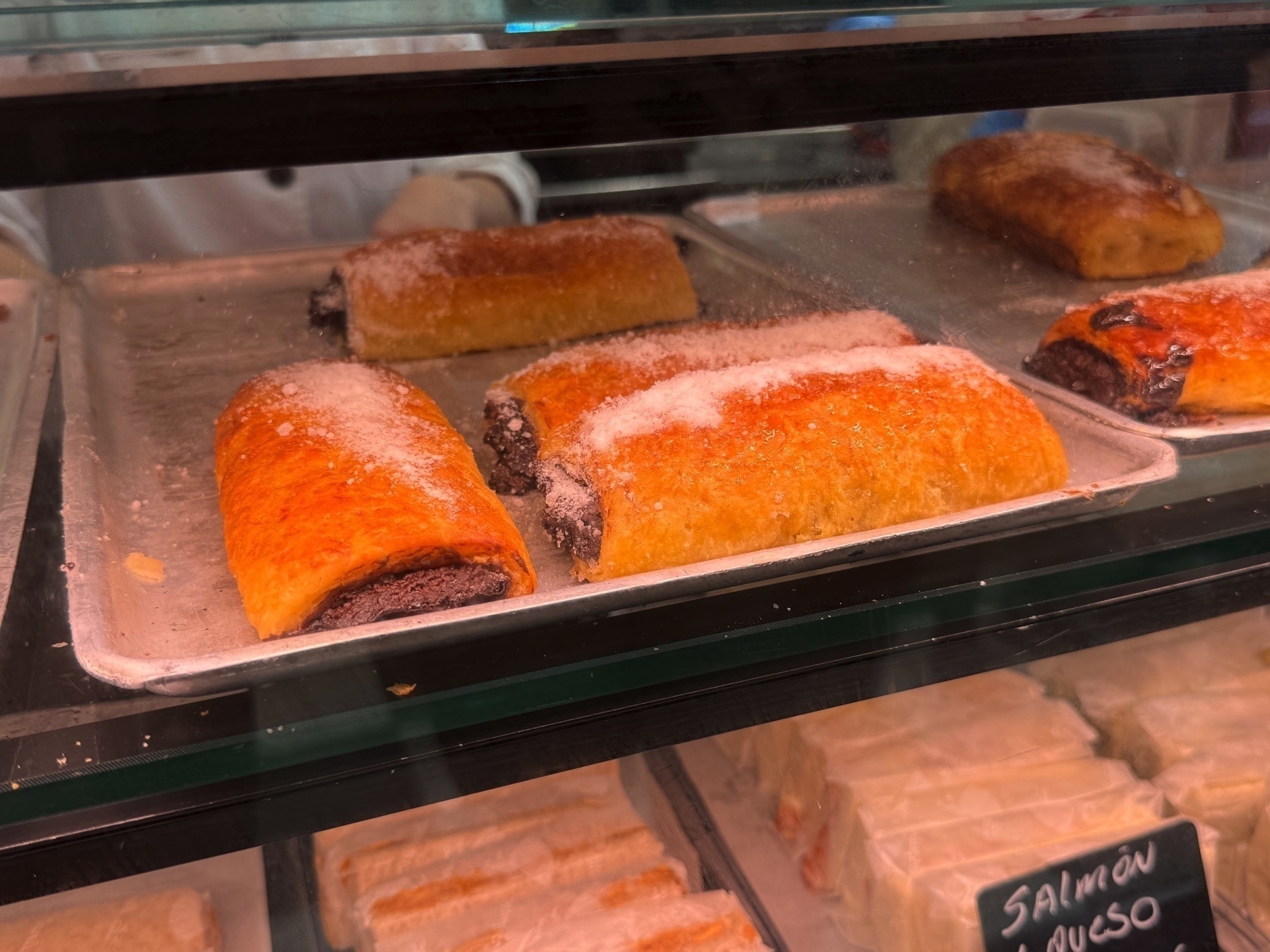 A bakery display case with sugar-dusted pastries filled with chocolate cream, their golden, flaky crusts slightly glossy under the lighting. The neat rows and rich filling peeking through the sides make these treats visually tempting and indulgent.