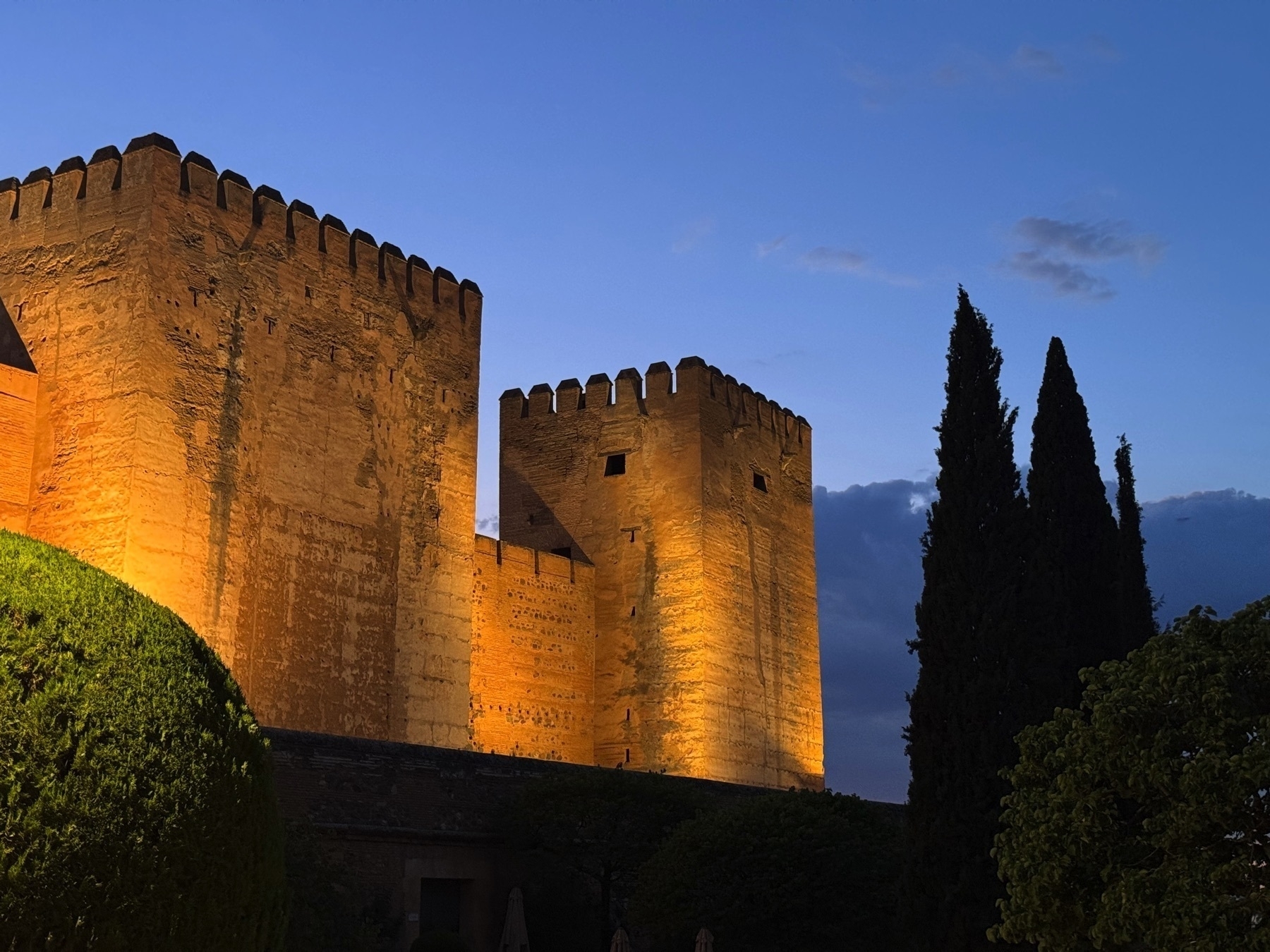 The Nasrid-era fortress walls and towers of the Alcazaba at the Alhambra glow with golden lighting at dusk, framed by manicured hedges and tall cypress trees under a clear evening sky.