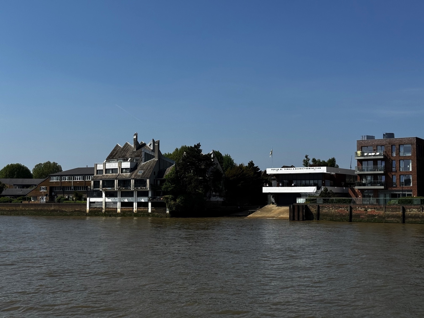 A riverside scene with modern and uniquely shaped buildings, including a sharply angled structure with multiple rooflines. A building labeled "Poplar, Blackwall and District Rowing Club" sits near the water’s edge, surrounded by trees and contemporary residences.