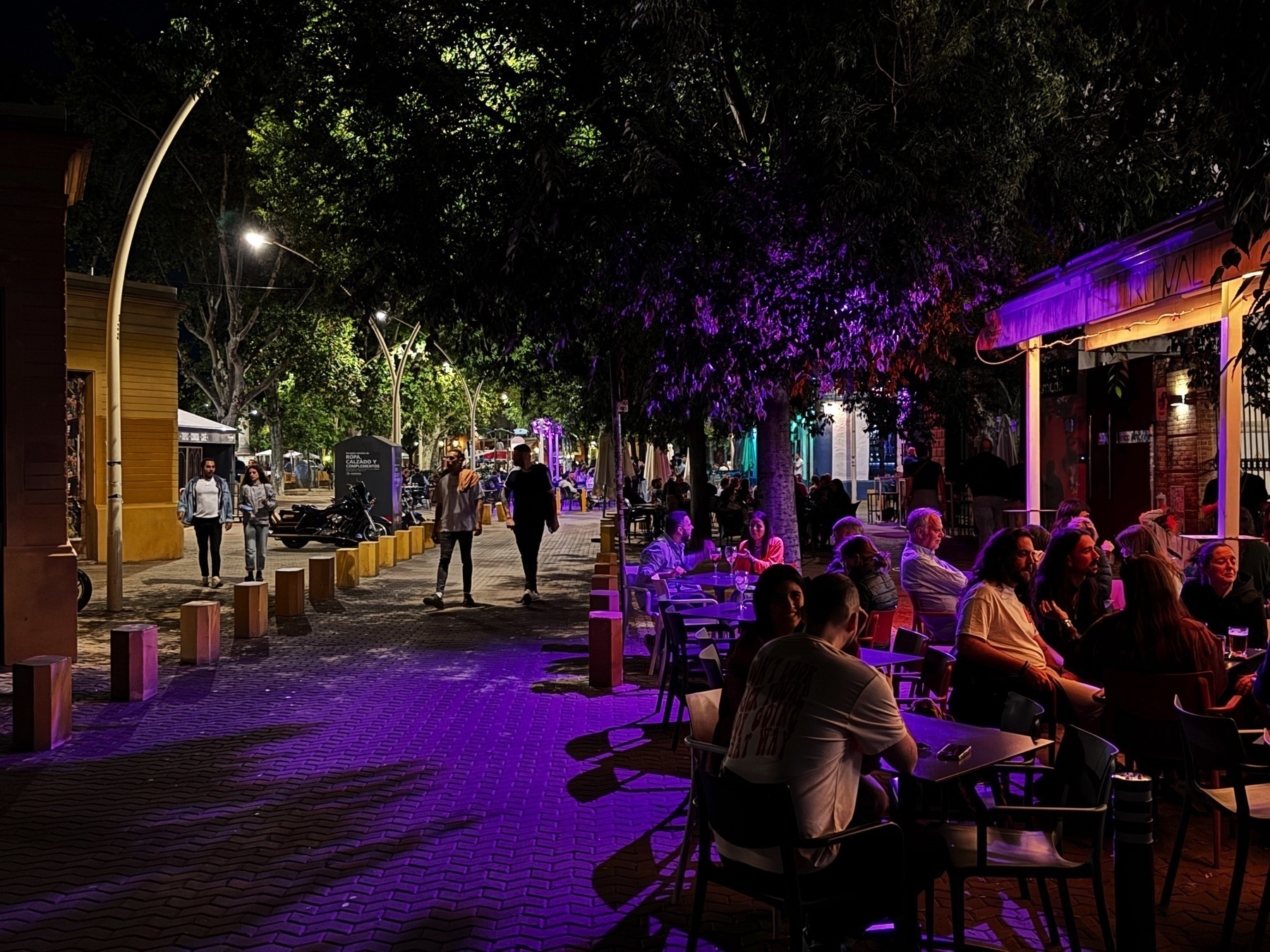 People enjoy a vibrant evening at an outdoor café on a pedestrian street, surrounded by trees lit with colorful purple and yellow lighting. Diners chat at small tables under a canopy, while others stroll along the walkway lined with modern streetlights and bollards.