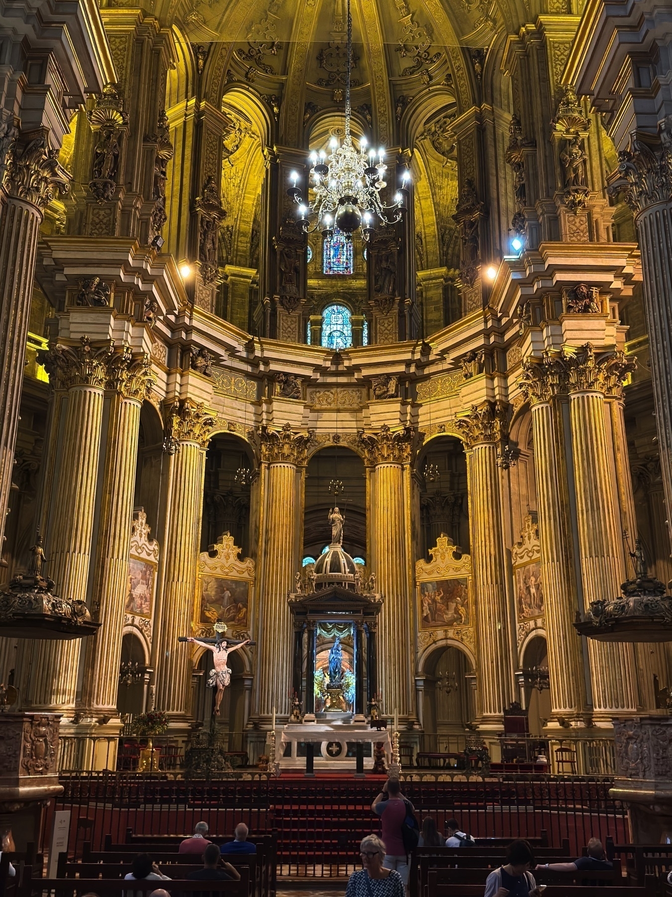 Interior view of the grand altar inside the Málaga Cathedral, featuring soaring Corinthian columns, gilded architectural details, a richly decorated vaulted ceiling, and a large statue of the Virgin Mary surrounded by intricate carvings and stained glass windows. Visitors are seated quietly in the pews, admiring the awe-inspiring beauty.