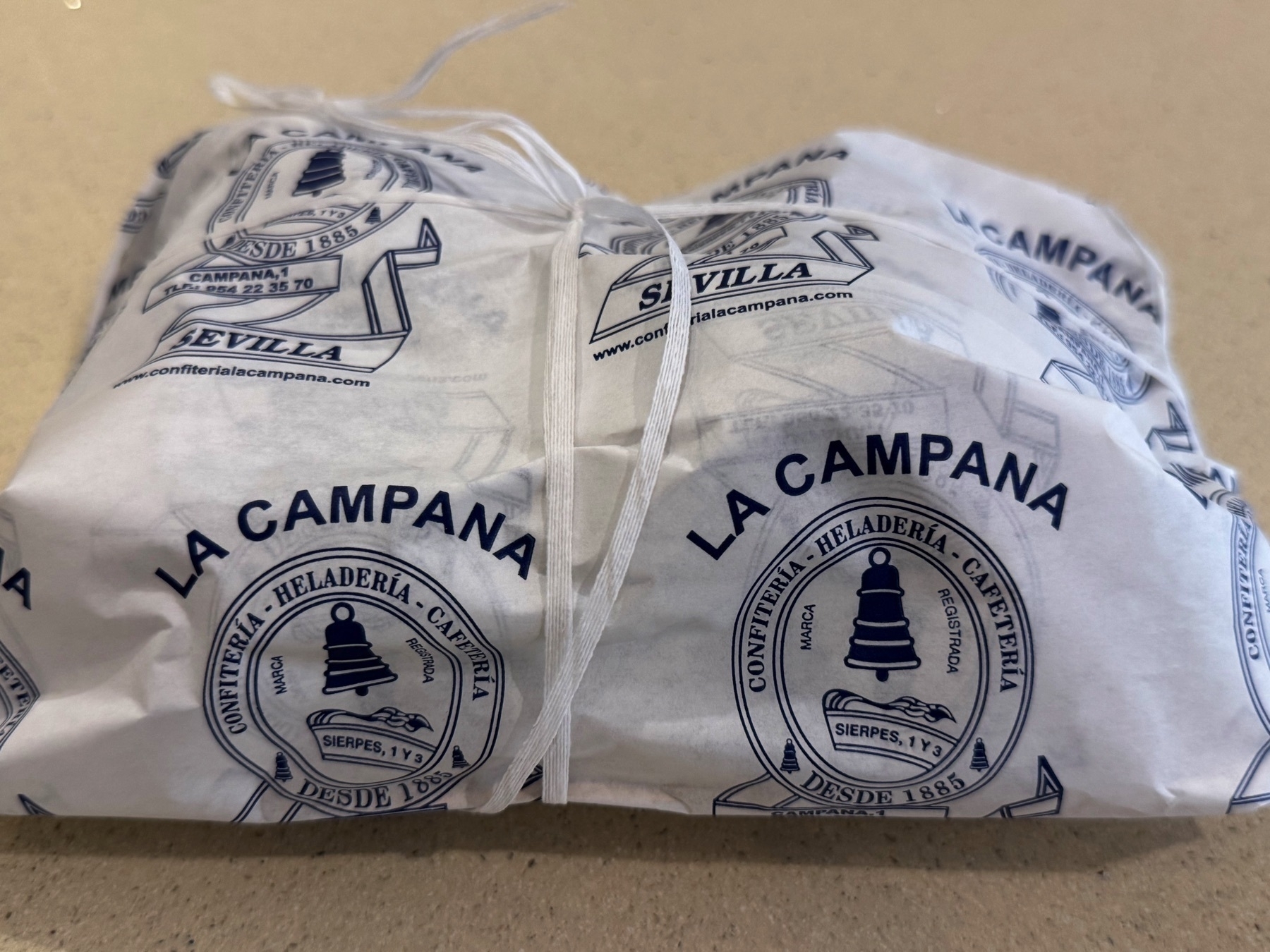 A neatly wrapped paper package from La Campana, a historic pastry shop in Seville, features blue printed branding with a bell logo and decorative script. The package is tied with white string and rests on a beige countertop, suggesting a freshly purchased dessert or pastry.