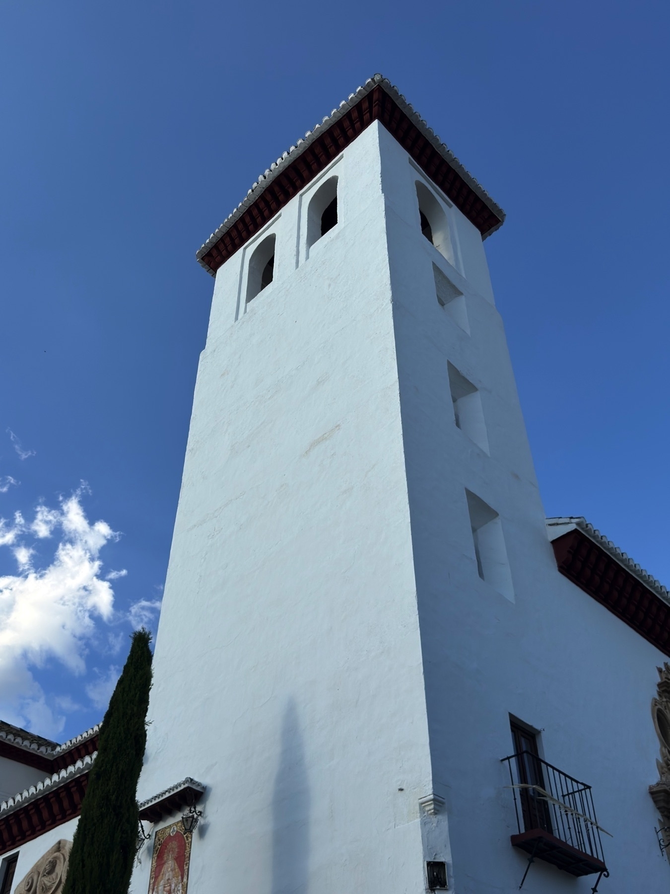 Upward view of a tall white bell tower with small arched openings, capped by a red-tiled roof. The clean lines and bright stucco façade contrast sharply against the deep blue sky.