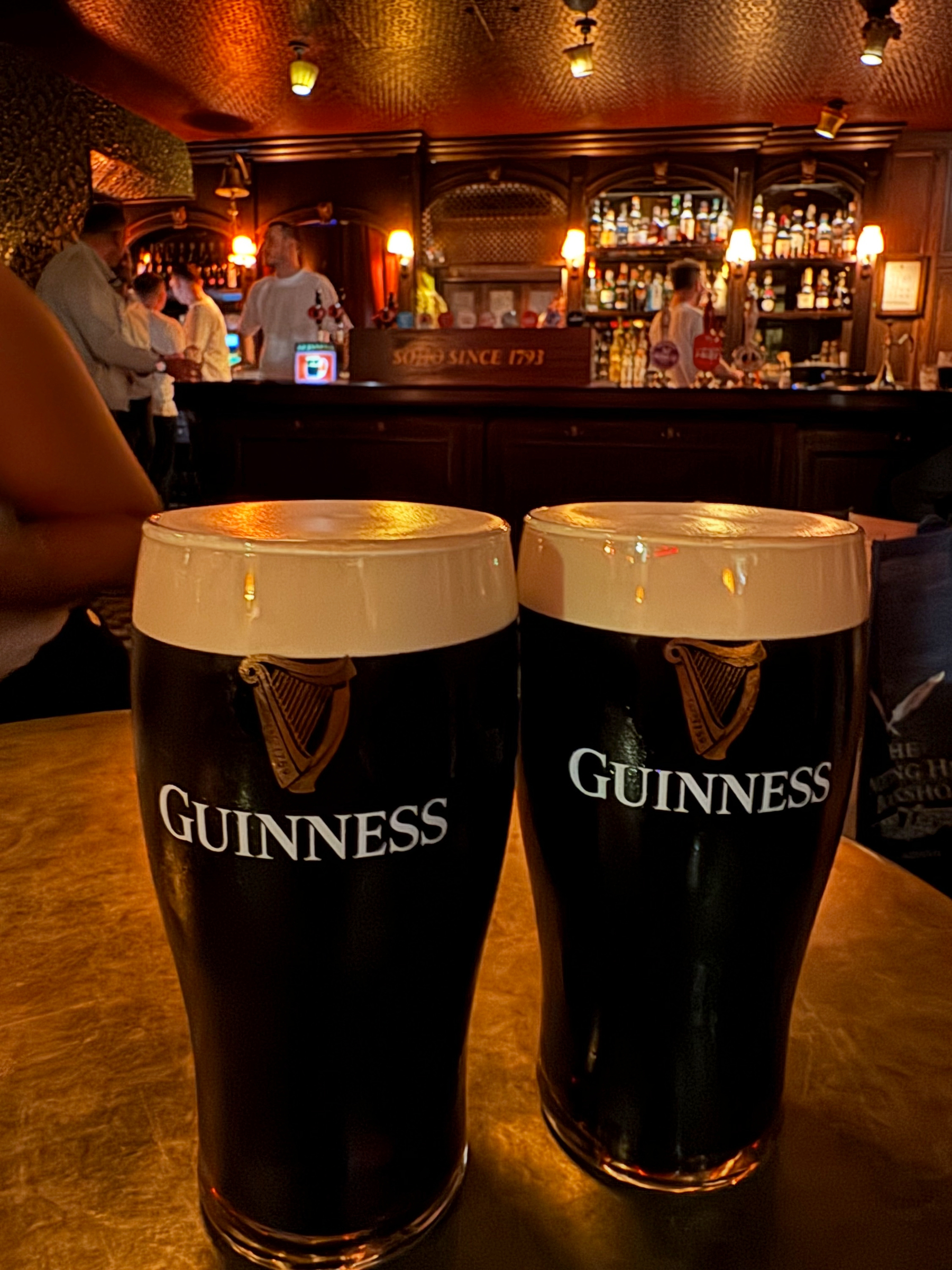 Two freshly poured pints of Guinness with thick, creamy heads sit on a small round table, the dark stout glowing slightly under warm pub lighting, with bartenders and a polished back bar of bottles softly blurred behind them. A sign reads “Soho since 1793.”