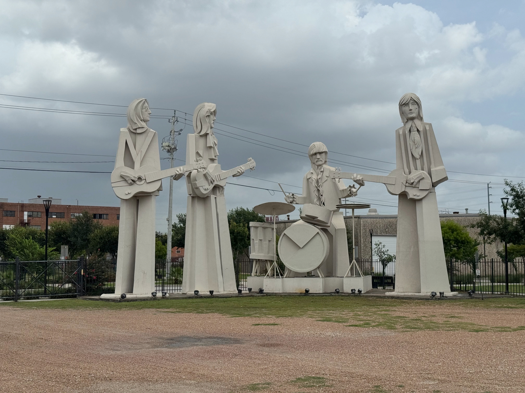 Four stylized stone statues of the Beatles standing in a line outdoors, each holding an instrument while a drummer sits at a kit, set against grass, industrial buildings, and an overcast sky.