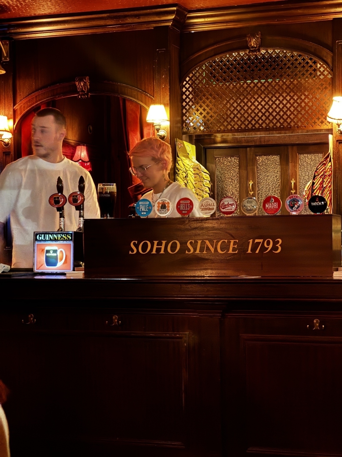 Two bartenders work behind a dark wooden bar with ornate decor, multiple branded beer taps, and a Soho Since 1793 sign under warm ambient lighting.