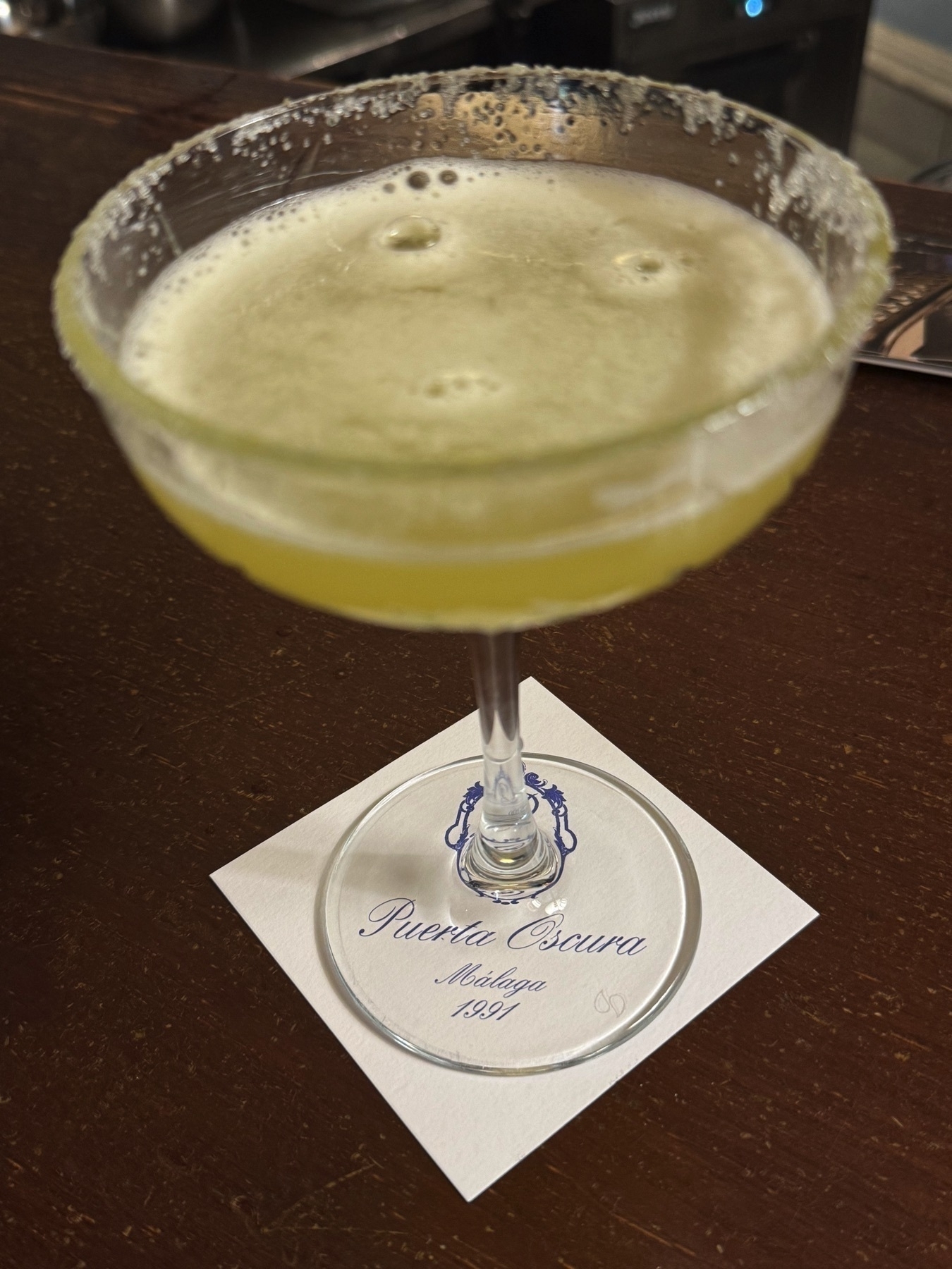 A pale yellow Margarita served in a coupe glass with a sugared rim, resting on a coaster from Puerta Oscura. The foamy surface and elegant presentation suggest a carefully crafted cocktail.