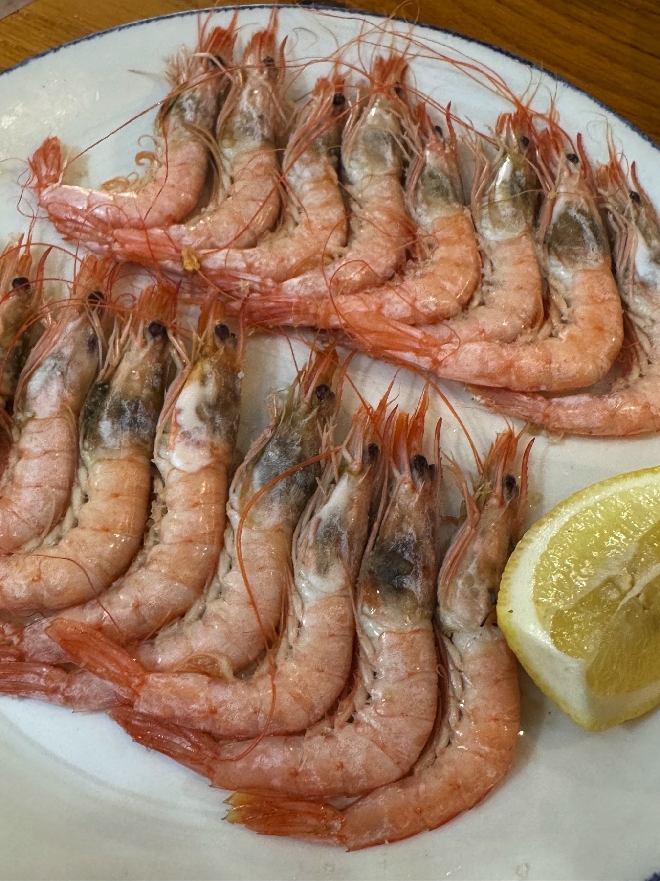 A chilled plate of pink prawns neatly arranged in rows around a lemon wedge. Their glistening shells and slightly translucent appearance suggest freshness and a clean, briny flavor, typical of expertly prepared cold seafood.