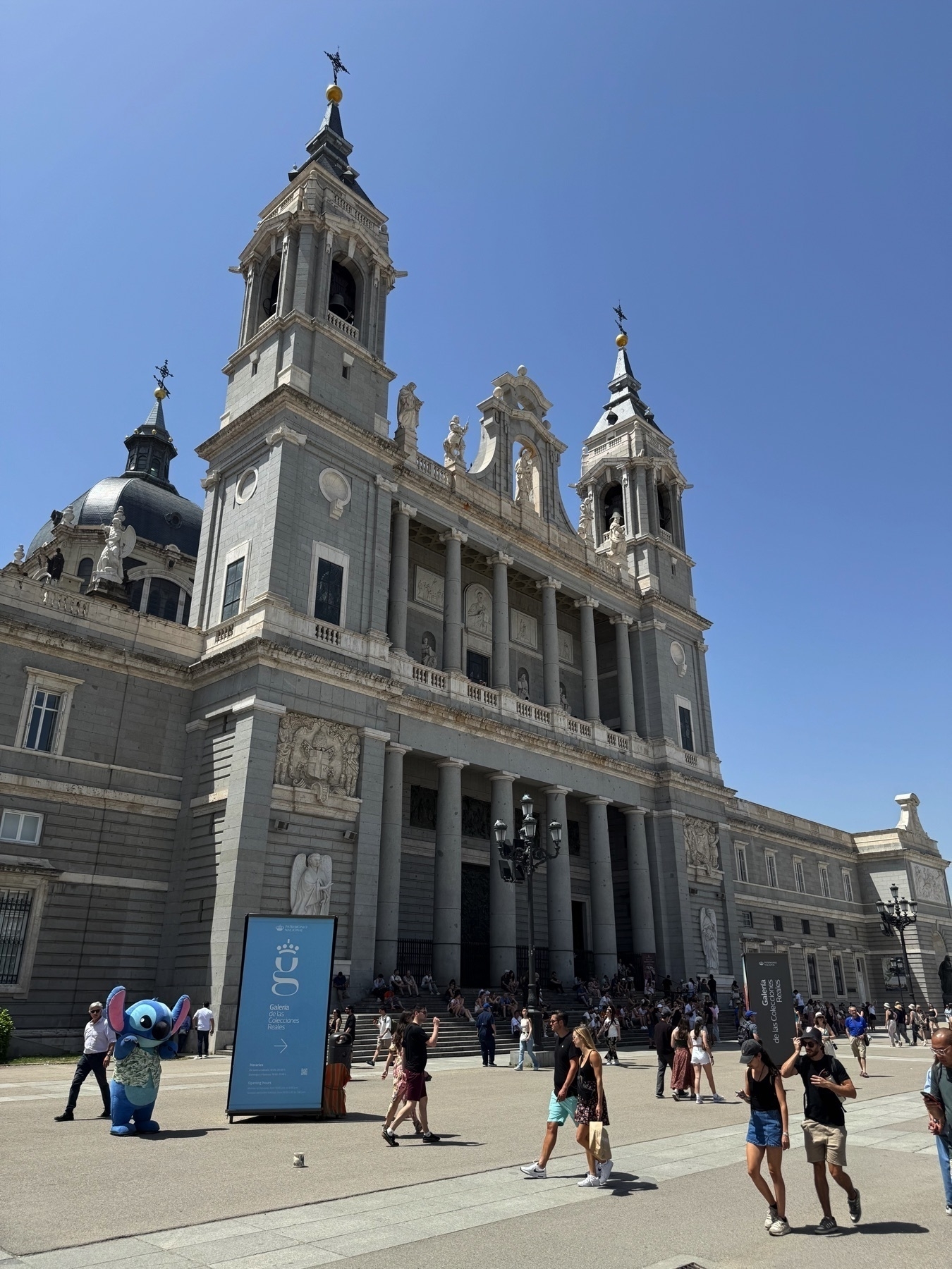 Lively street-level view of the Almudena Cathedral from an angle that showcases its detailed exterior, including baroque and neoclassical elements, multiple spires, and the central dome. Crowds gather at the cathedral’s entrance, and a person in a Stitch costume adds a playful touch near signage for the museum. The vibrant activity and crisp shadows emphasize the warm, sunny day.