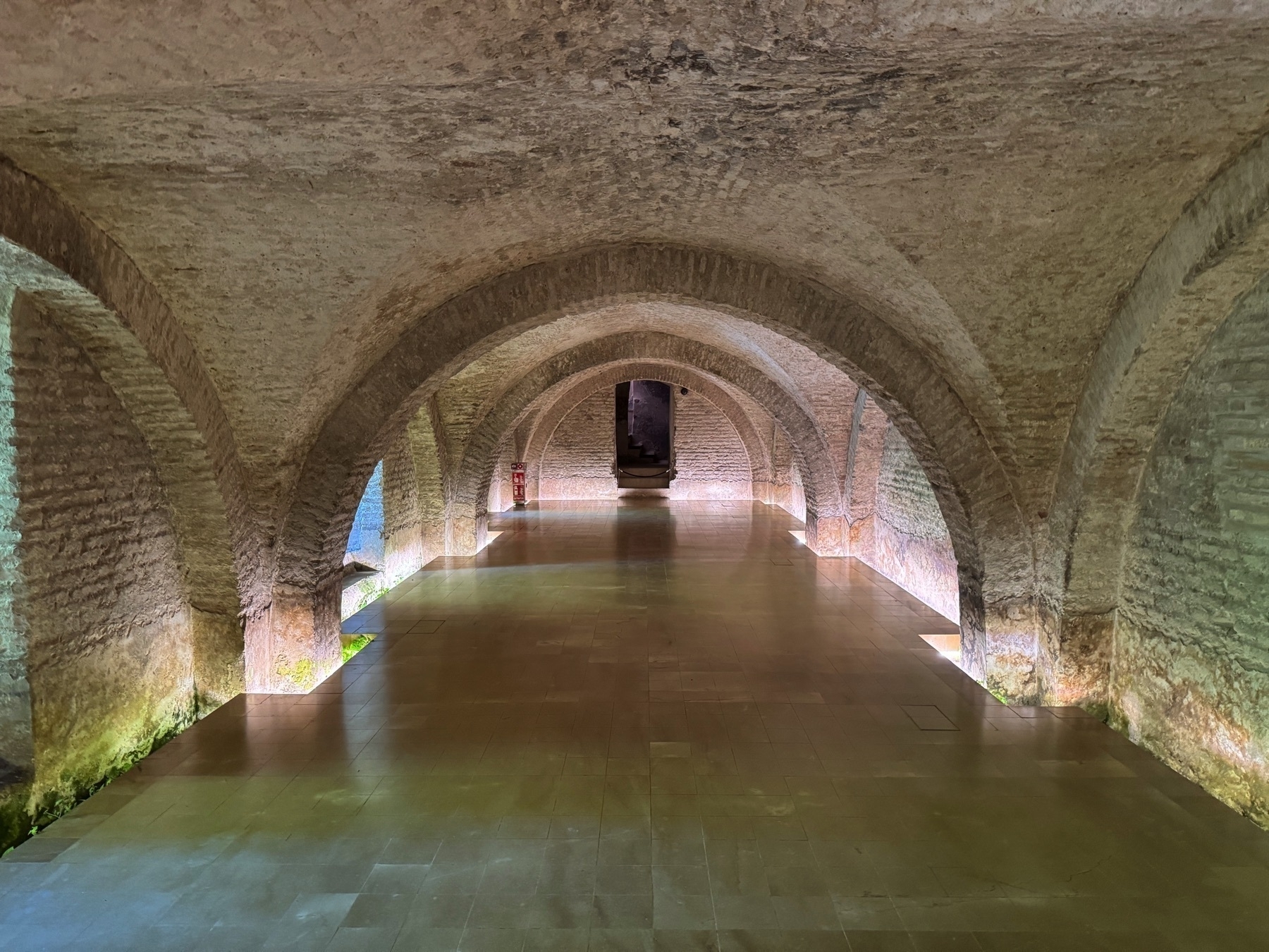 The Baths of Doña María de Padilla, a vaulted underground chamber with brick arches and a shallow reflecting pool. The space is softly lit by natural light, emphasizing its symmetry and Gothic architectural form.