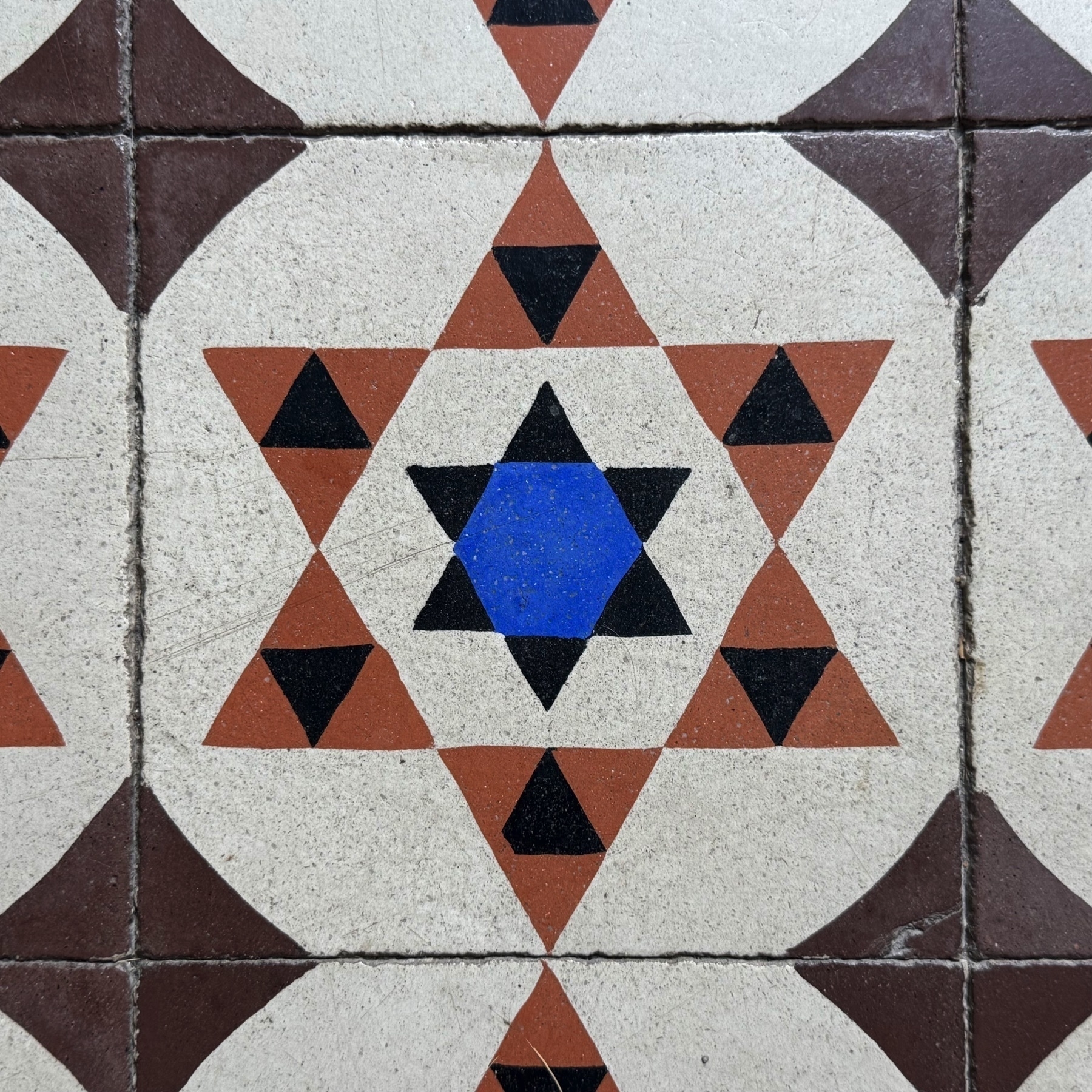 Close-up of decorative floor tiles featuring a geometric pattern composed of interlocking stars. A central blue hexagon is surrounded by black and brown triangles forming a six-pointed star, repeated across the surface. The symmetry and bold color contrast create a visually striking and intricate design.