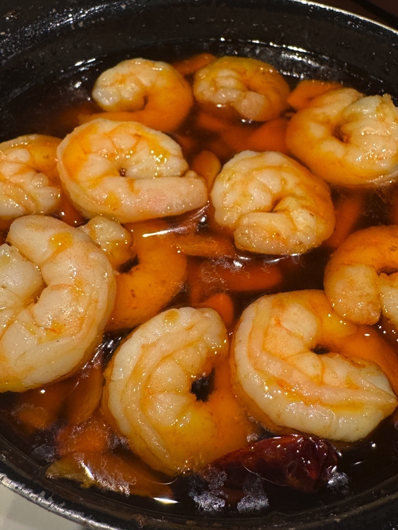 Plump shrimp sizzling in hot olive oil with visible pieces of garlic and dried red chili peppers, served in a small black cazuela that captures the bubbling oil and rich golden tones of the dish.