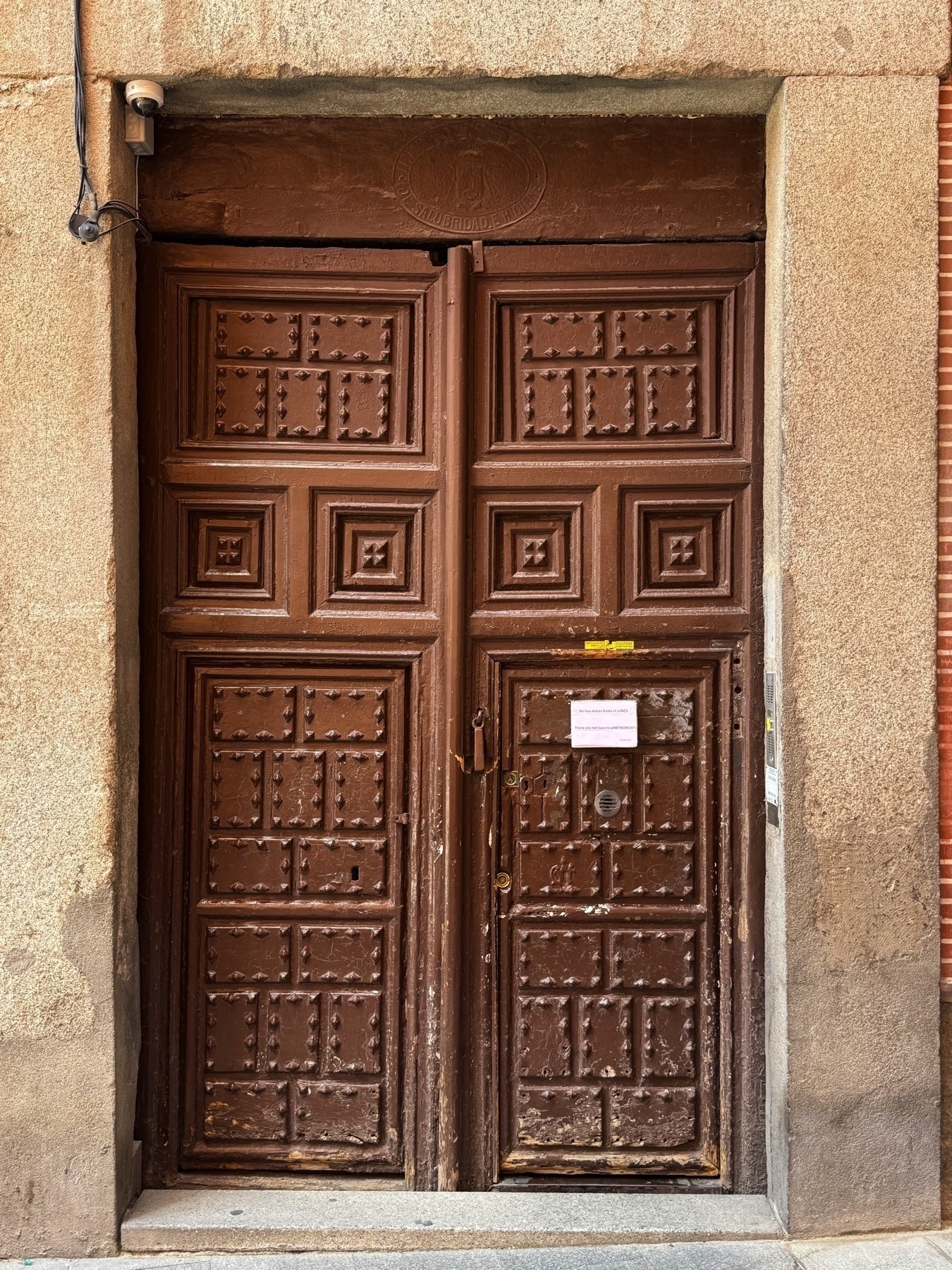 Full view of a tall, dark brown wooden door set in a stone frame, featuring a pattern of square and diamond-studded panels. A small laminated notice is posted on one side of the door. A security camera is mounted above, and a “Mind your head” warning label is attached to the door lintel. The weathered surface and heavy design convey a sense of historical weight.