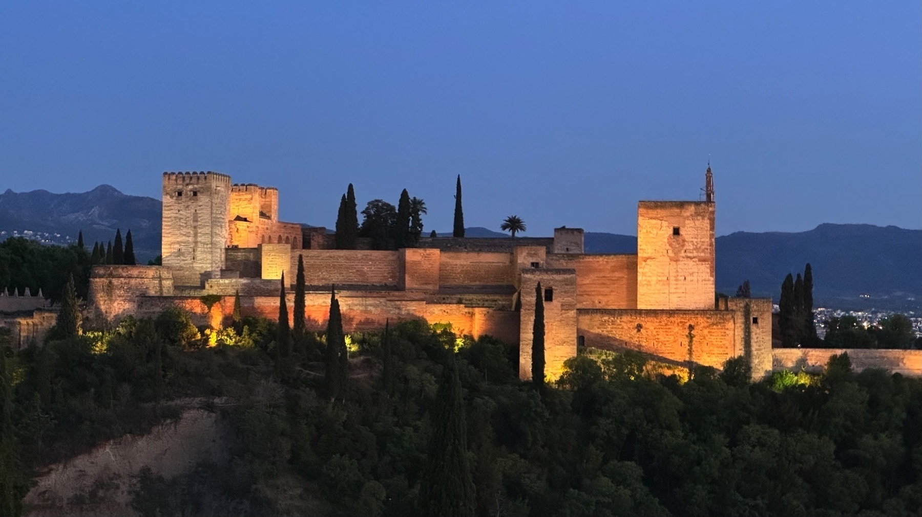The Alhambra illuminated at dusk, with golden light highlighting the reddish stone walls and towers against a deepening blue sky. Cypress trees line the outer perimeter of the fortress, and the silhouettes of distant mountains frame the background.