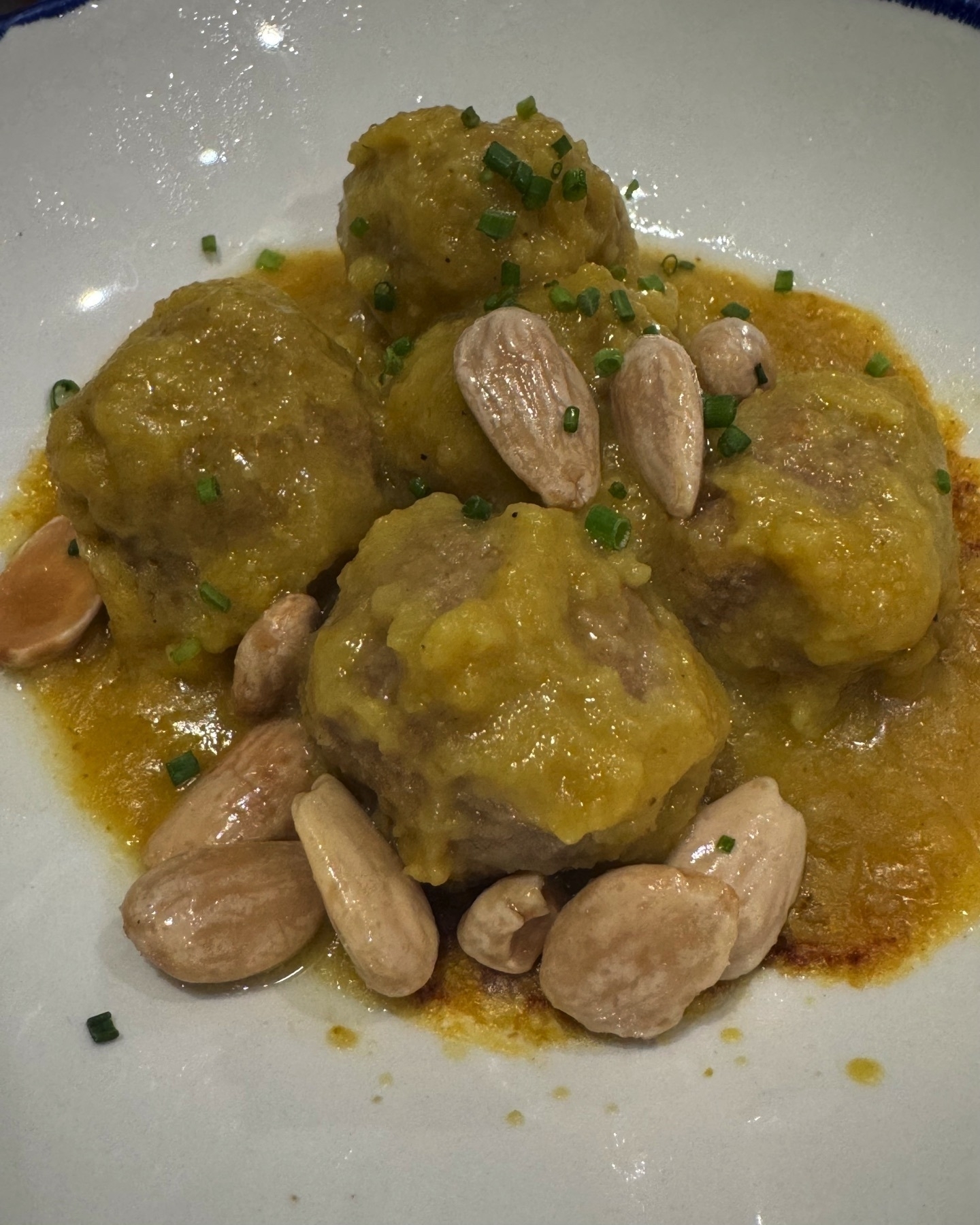 Tender meatballs in a thick golden sauce, garnished with blanched whole almonds and finely chopped chives, presented on a plain white plate that highlights the warm tones of the dish.