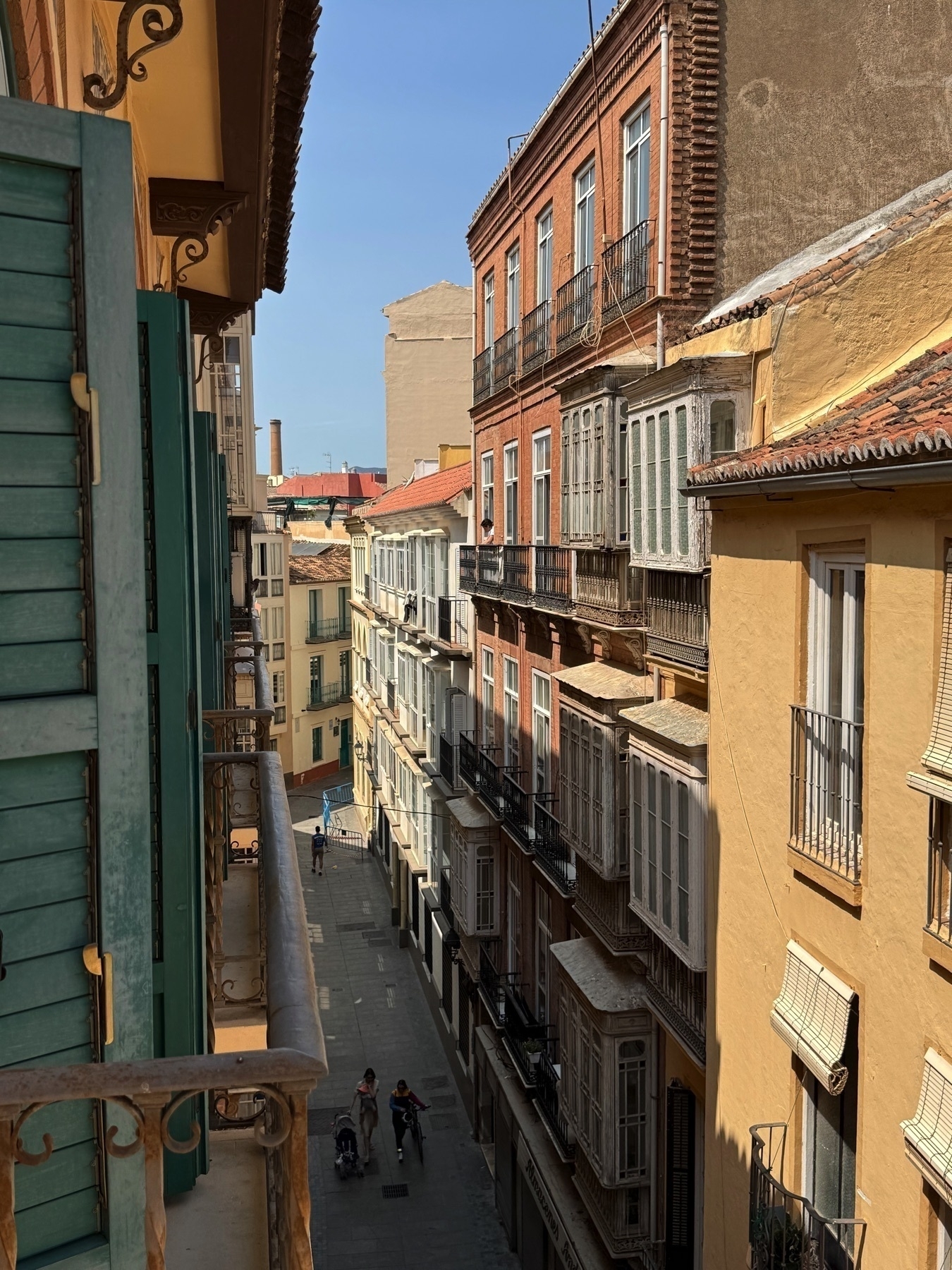 Downward view from a balcony onto a narrow, shaded street lined with classic Andalusian buildings with iron railings and wooden shutters. The perspective leads the eye through the pedestrian path below, where a few people stroll or push strollers. Soft light and pastel-colored walls give the image a warm, inviting feel.