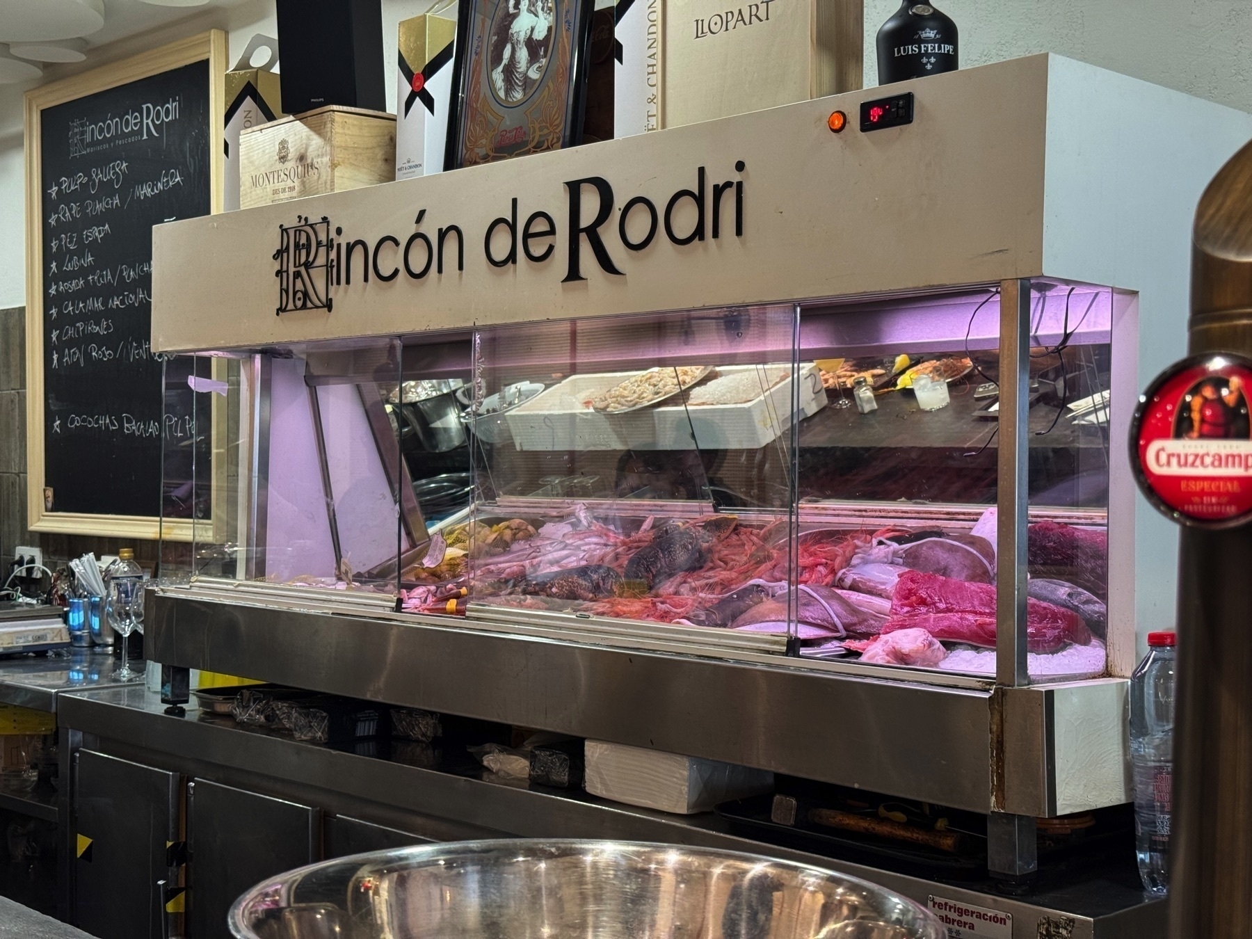 A fresh seafood display behind glass at the bar, with various fish, shellfish, and crustaceans laid on crushed ice. The counter is lit from above, giving the seafood a vibrant, just-caught look, while the metallic surfaces and stacked wine boxes create an inviting, market-style ambiance.