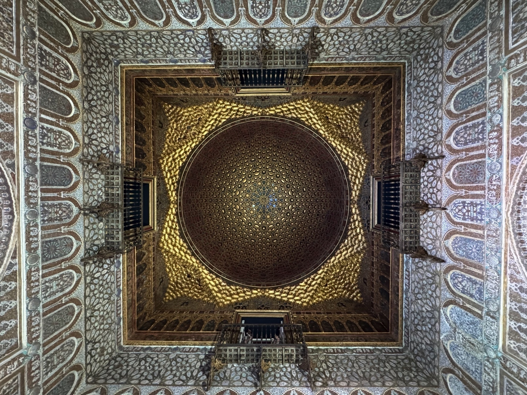 Ceiling of the Hall of Ambassadors, featuring a gilded wooden dome in the _mocarabe_ style. The interlaced star pattern and carved woodwork are surrounded by highly decorated walls with repeating plaster motifs and projecting balconies.