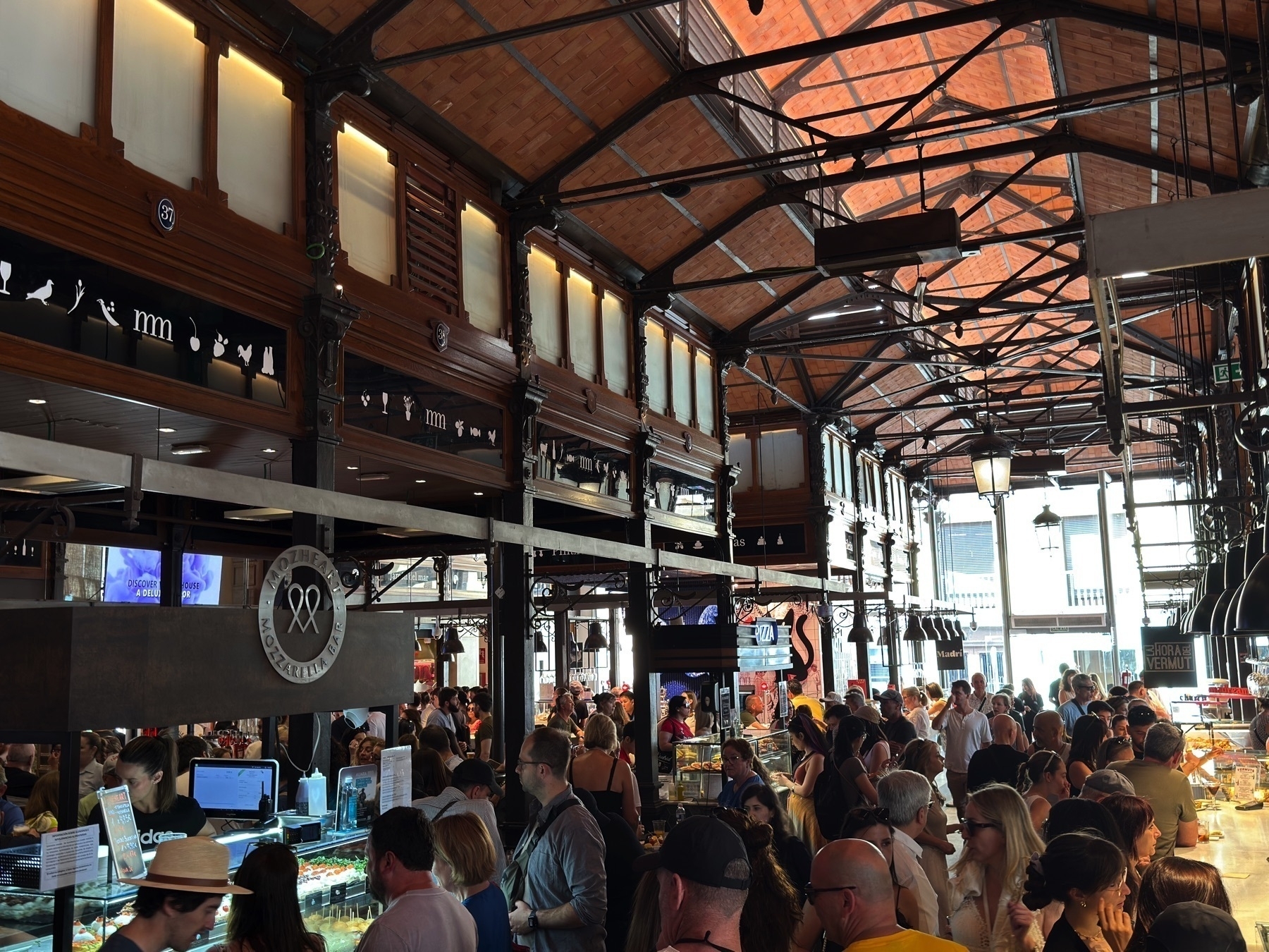 Bustling interior of the market with a crowd of visitors navigating food stalls beneath a vaulted ceiling with exposed iron trusses and terracotta tiles. The warm ambient light and wooden stall facades create a cozy yet vibrant atmosphere. Hanging signage and black pendant lamps help define each vendor space, with people lining up for tapas and drinks.