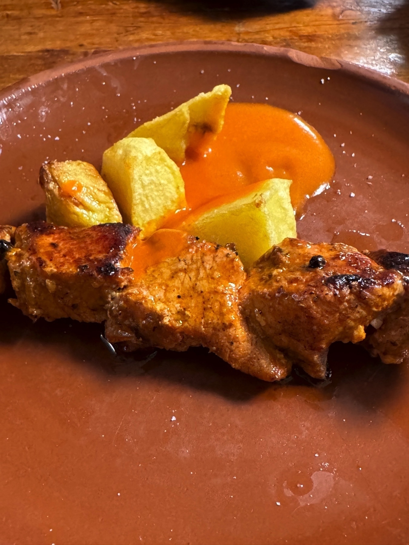 A terracotta plate with a skewer of grilled marinated pork, slightly charred and coated in a vibrant red sauce. Alongside are a few chunky fried potatoes, partially covered in the same sauce, creating a simple yet flavorful presentation.