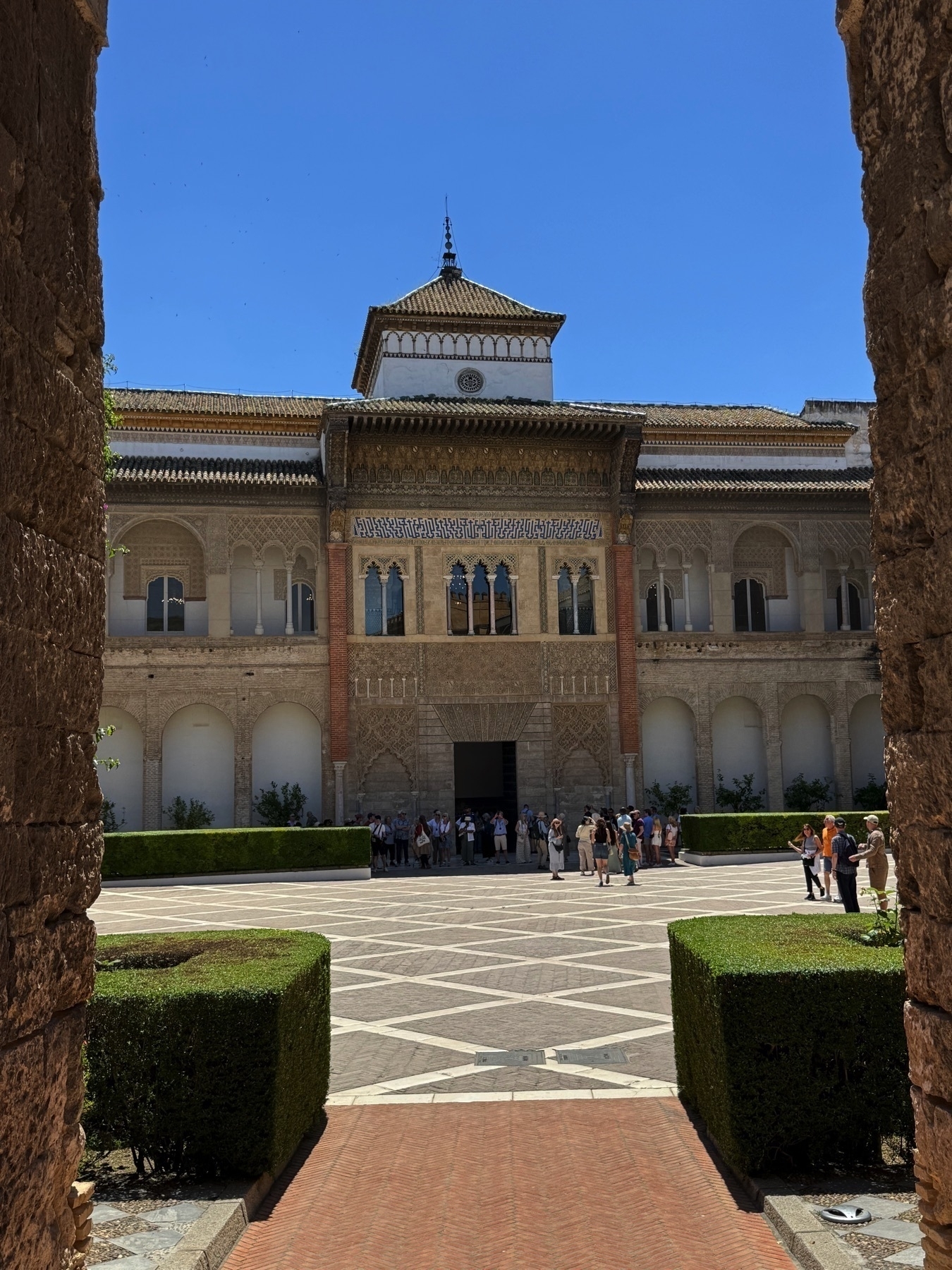 View of the courtyard in front of the Palacio del Rey Don Pedro, showing Islamic-style architecture with a large horseshoe arch, geometric plaster decoration, and a tile roof. Visitors stand before the building, surrounded by hedges and patterned paving.