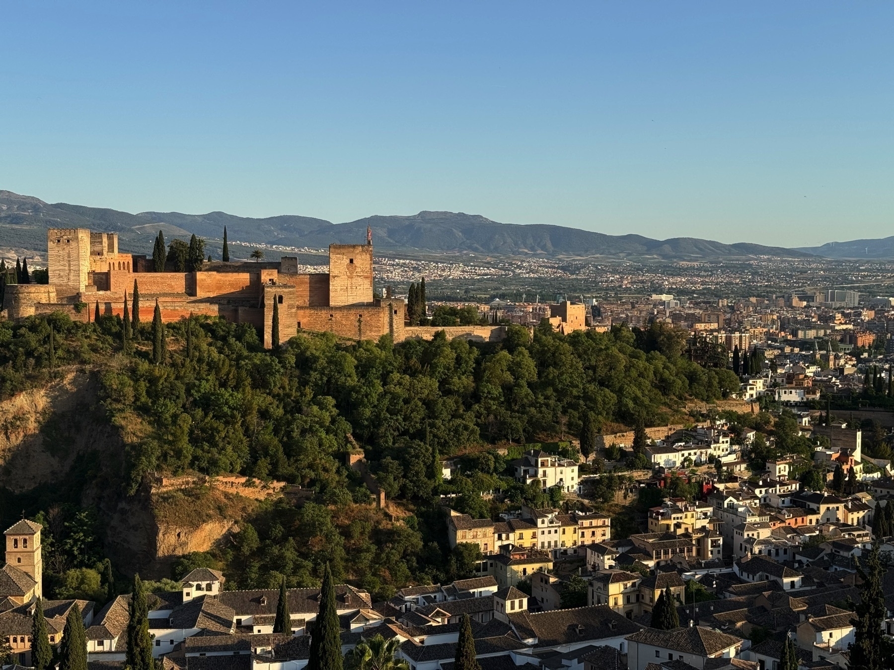 Wide view of the Alhambra in early evening light, perched above the Albaicín neighborhood of Granada. The terracotta roofs of whitewashed houses spread out in the foreground, with the fortress standing prominently above dense green vegetation and backed by distant mountains.
