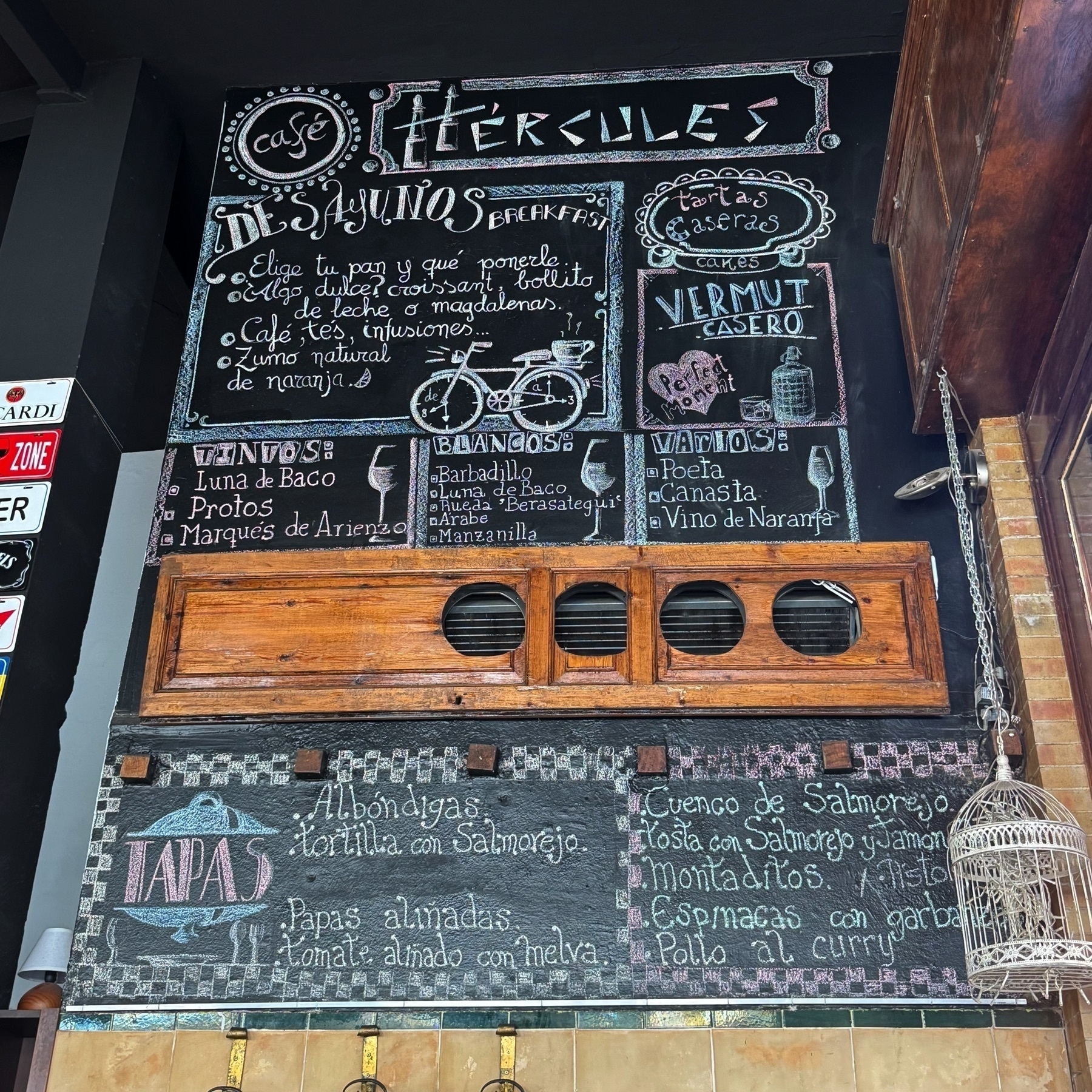 A large chalkboard wall inside Café Hércules displays handwritten Spanish menu items including tapas, vermouth, wines, and breakfast choices, surrounded by hand-drawn illustrations like a bicycle and teacups. The colorful, whimsical typography and hanging birdcage contribute to the eclectic, creative vibe of the café.