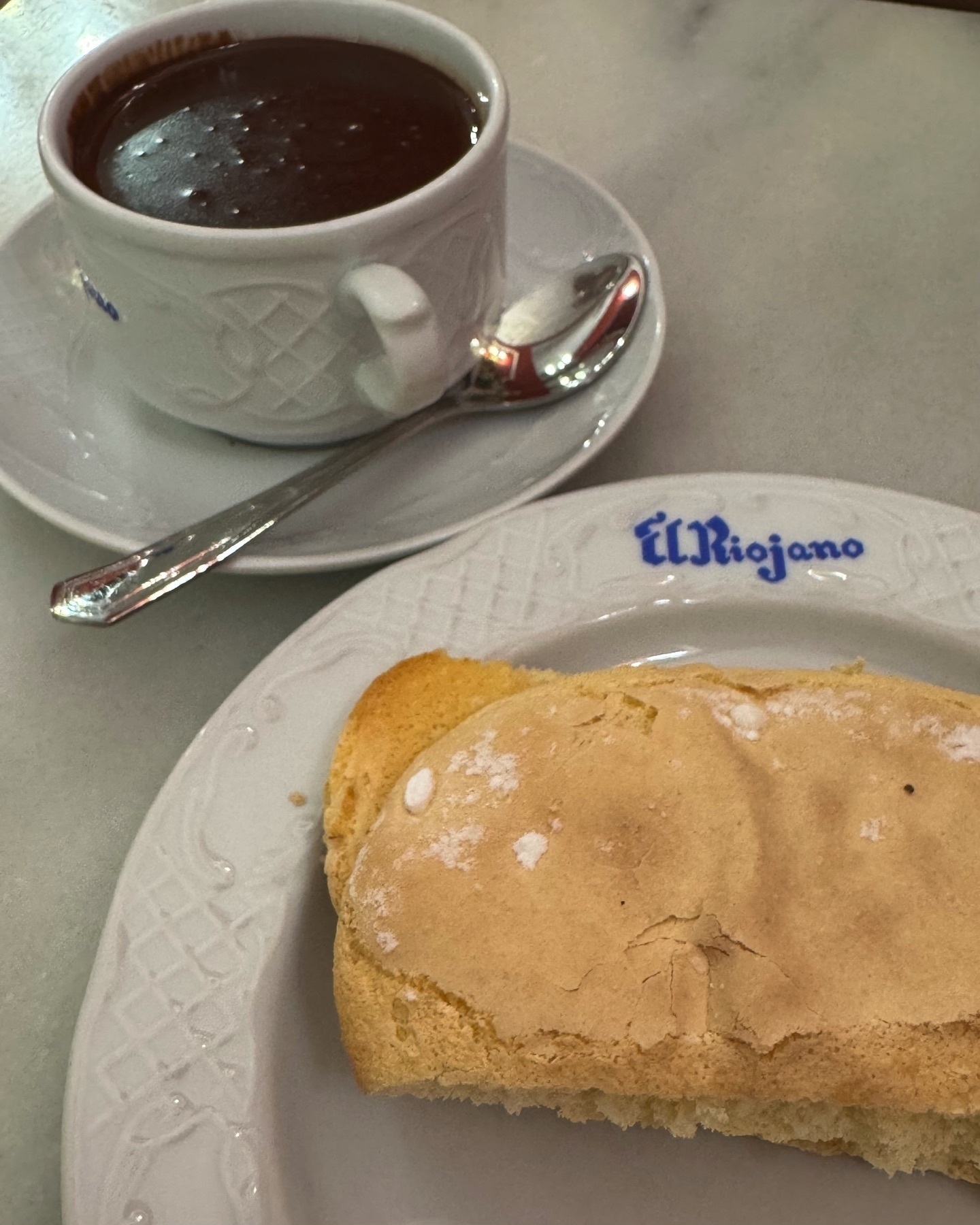 A thick cup of Spanish hot chocolate sits on a white saucer with an ornate handle and a silver spoon, paired with a soft, golden sponge cake dusted lightly with powdered sugar. Both are served on fine white porcelain dishes embossed with patterns and marked with the name “El Riojano” in cobalt blue, evoking a sense of elegance and tradition. The chocolate’s glossy surface and dense texture contrast invitingly with the tender, airy look of the cake.