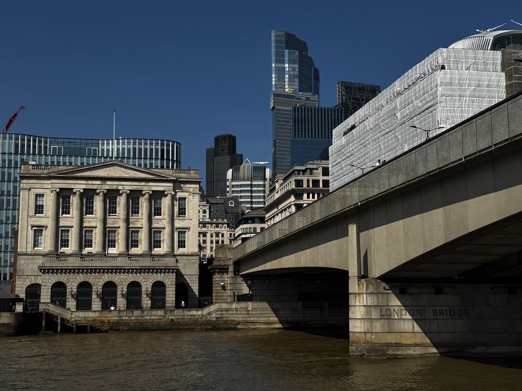 A view of London Bridge with its name inscribed on the stone base, next to a neoclassical building with columns. Behind them, a dense cluster of glass and steel skyscrapers contrasts with the older architecture near the riverbank.