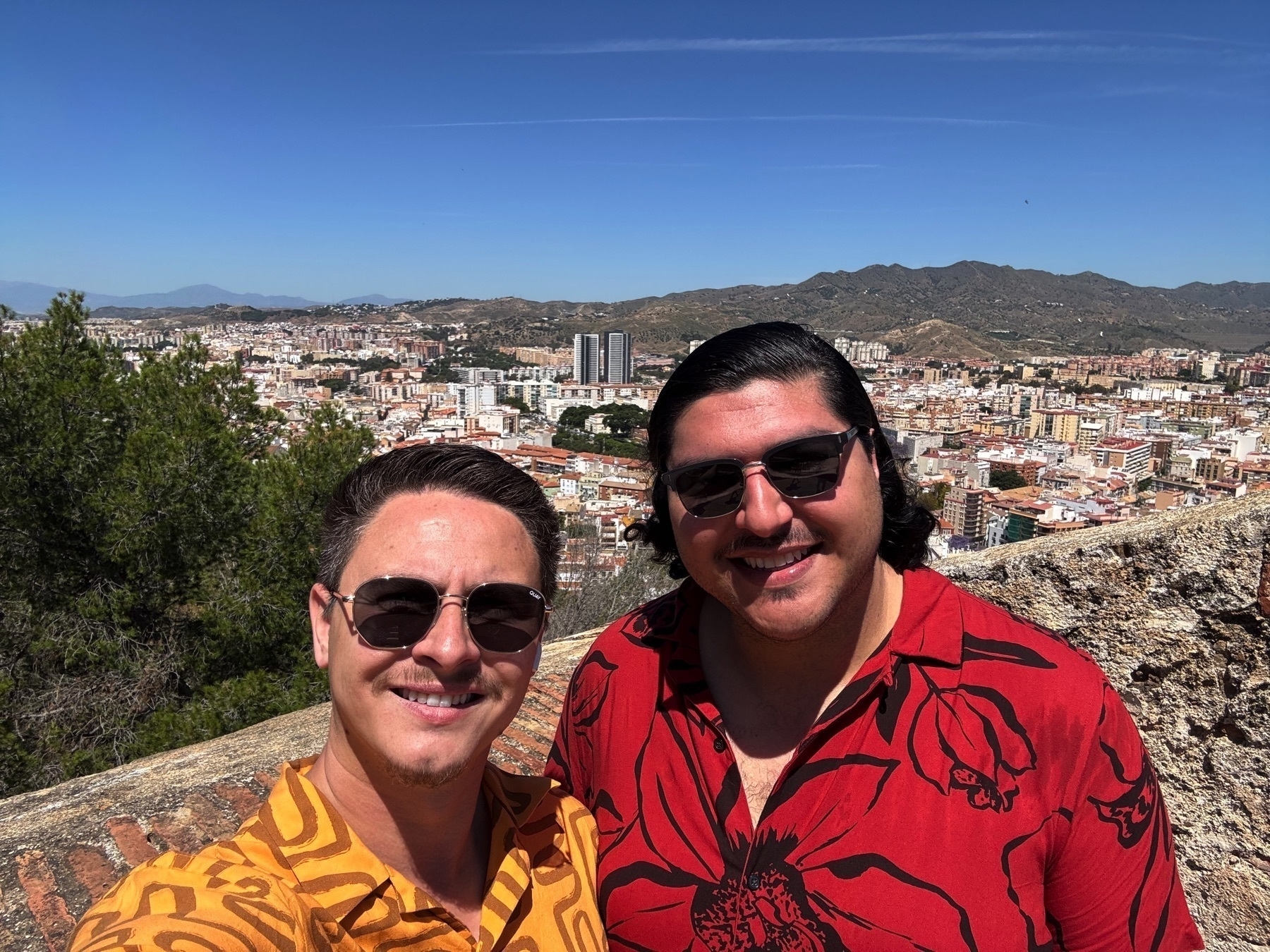 Two men smiling for a selfie with a panoramic view of Málaga in the background. They are standing on a stone terrace lined with bricks and trees, with the city and surrounding hills stretching into the distance under a clear blue sky. Both wear patterned shirts and sunglasses.