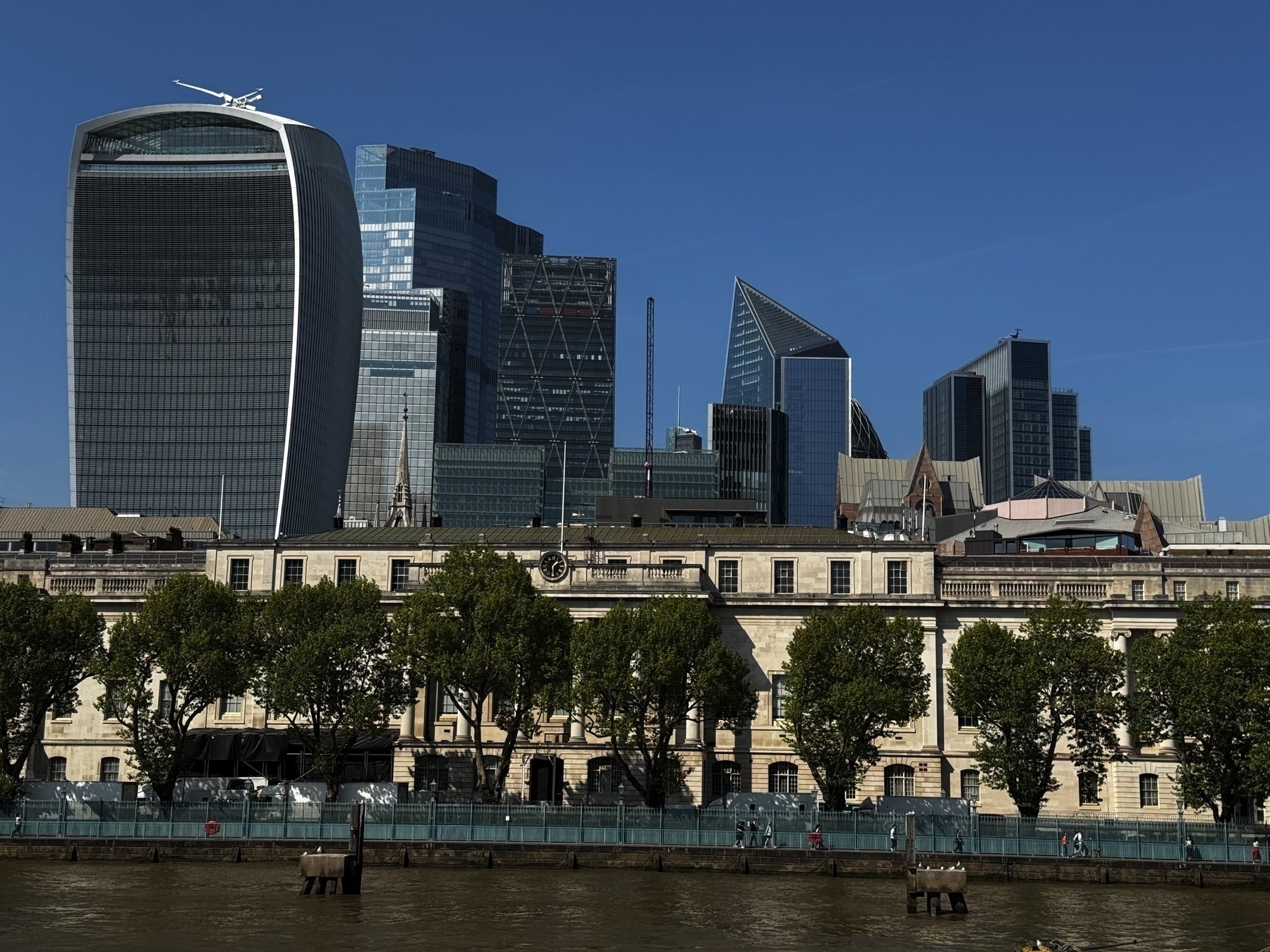 A cityscape view of central London from across the Thames, showcasing the curved façade of the Walkie-Talkie building alongside several modern glass skyscrapers rising behind a historic stone building and a row of green trees.