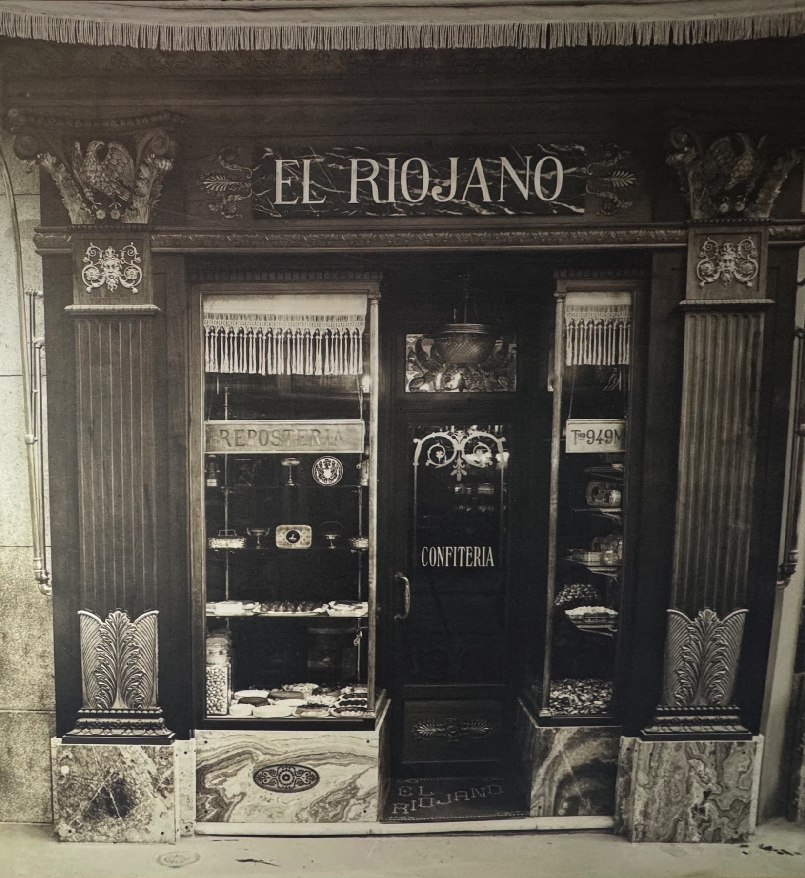 A vintage black-and-white photograph of a traditional storefront labeled “El Riojano,” featuring ornate classical columns and carved wooden detailing around a narrow entrance. Two window displays on either side show tiered trays of confections and pastries, while the words “Repostería” and “Confitería” above the door frame the scene. The nostalgic ambiance reflects old-world charm and a long-standing legacy of fine pastry-making.