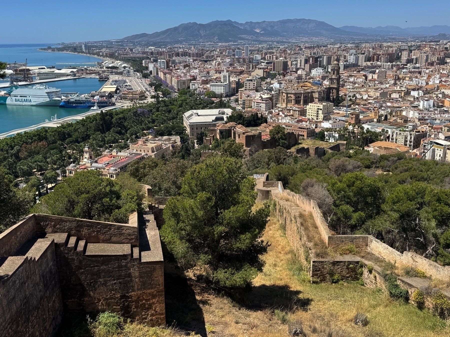 View from the Gibralfaro showing the historic and modern cityscape of Málaga, with the port, a large Balearia ferry, and the iconic Málaga Cathedral visible among dense urban buildings. The Mediterranean Sea and distant mountains create a scenic backdrop.