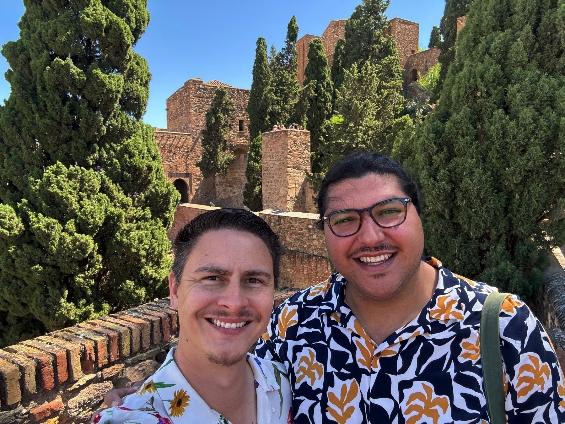 Two smiling partners take a selfie in front of the Alcazaba’s fortified walls, surrounded by dense greenery and tall cypress trees. The sun shines brightly, casting clear light over the historic stone towers.