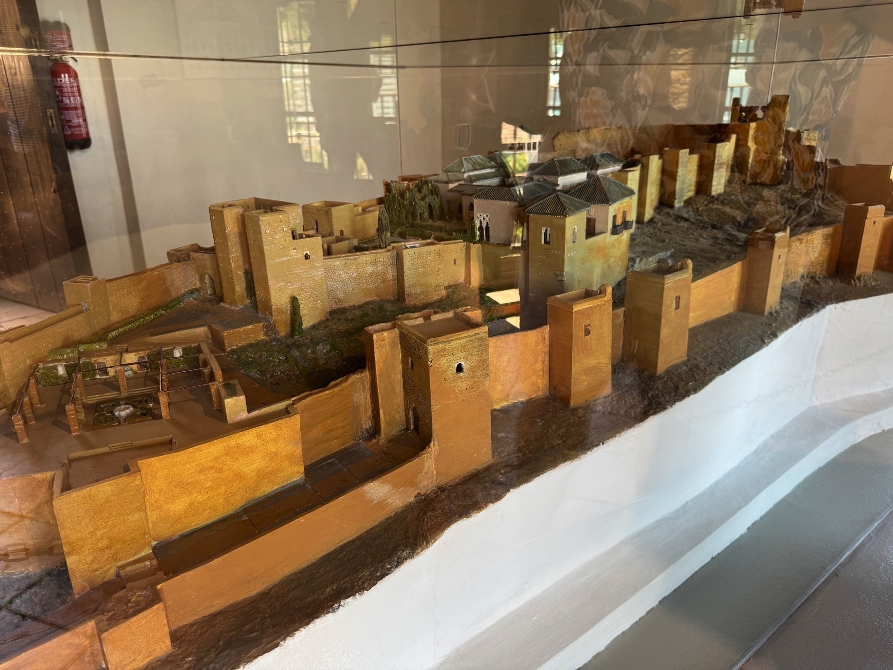 A detailed scale model of the Alcazaba displayed under glass, showing its full layout with fortified walls, towers, inner courtyards, and residential buildings. The miniature includes textures, vegetation, and terrain for realism.