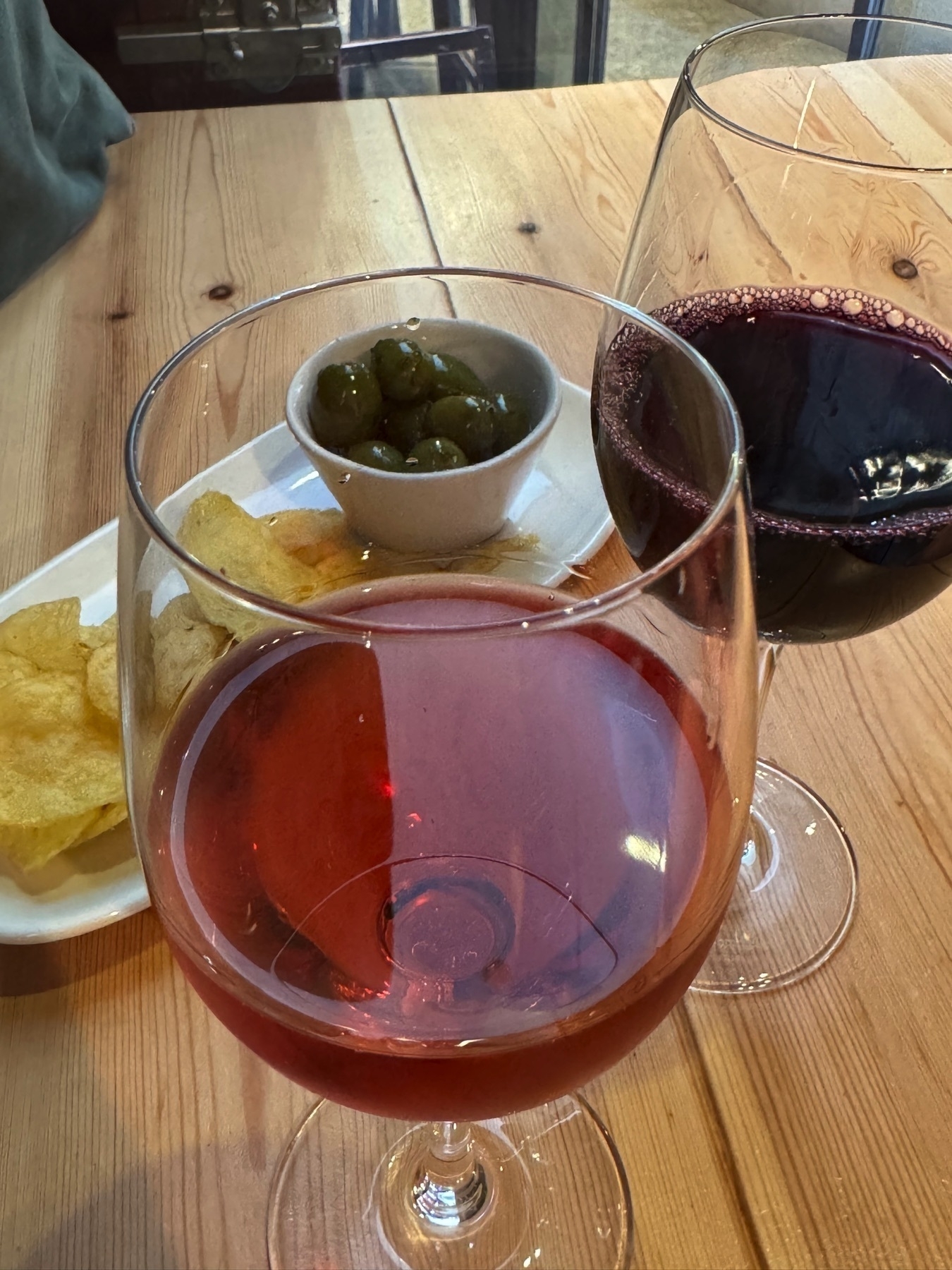 Two wine glasses—one filled with deep red wine, the other with a rosé—set on a light wooden table next to a small bowl of green olives and a few potato chips. The ambient light and casual tapas pairing evoke a relaxed, elegant dining atmosphere.