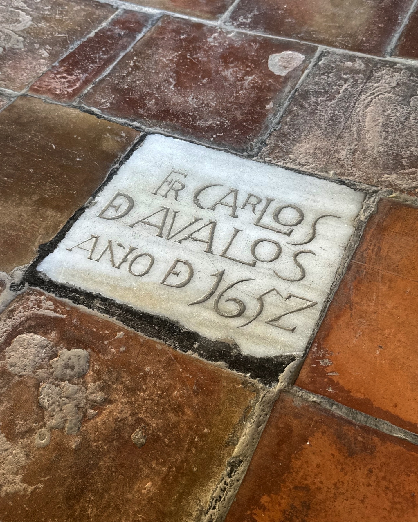 Marble grave marker inscribed with the name “Fr. Carlos Dávalos” and the year 1652, set among worn terracotta tiles. The engraved stone stands out against the earthy floor, evoking the long history of the site and its monastic inhabitants.