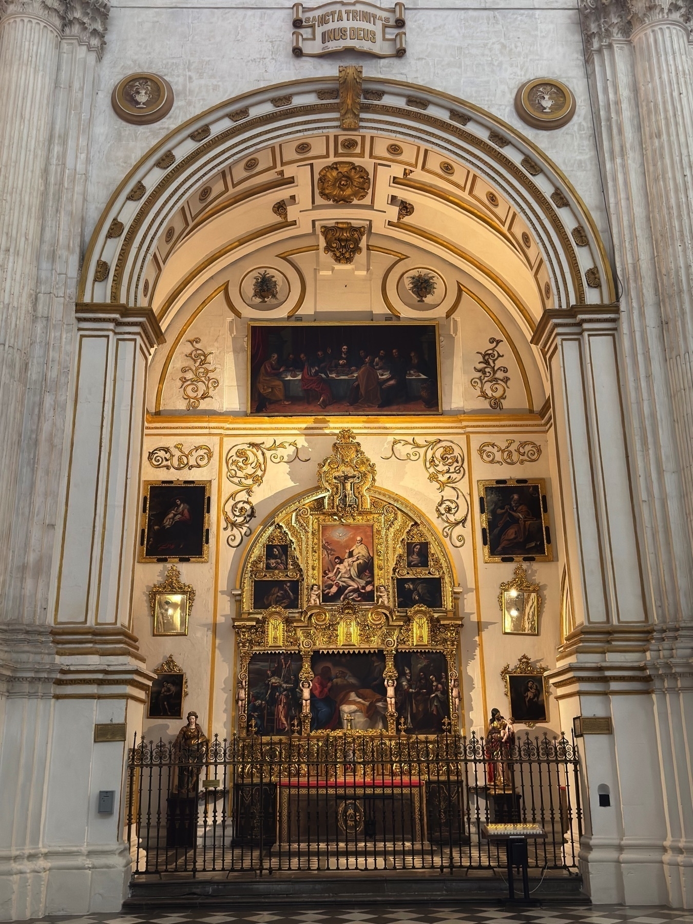 Side altar framed by rounded white columns and golden trim, featuring a gilded retablo adorned with religious paintings. A central crucifixion scene is flanked by smaller panels, each rich in color and emotion. The setting is contained behind a low metal gate, underscoring its sacred significance within the larger church interior.