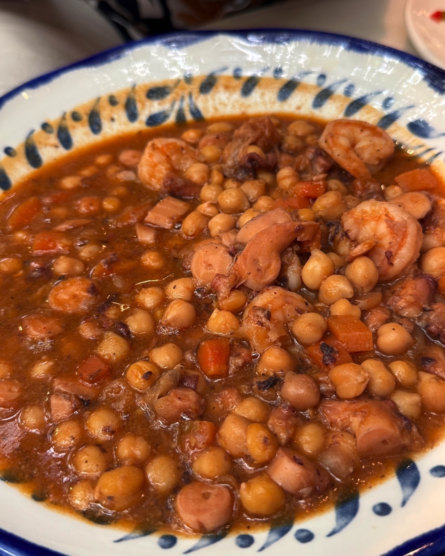 Hearty stew of chickpeas, shrimp, and pieces of octopus in a rich tomato-based broth, served in a deep blue-rimmed bowl with a rustic, home-cooked appearance.