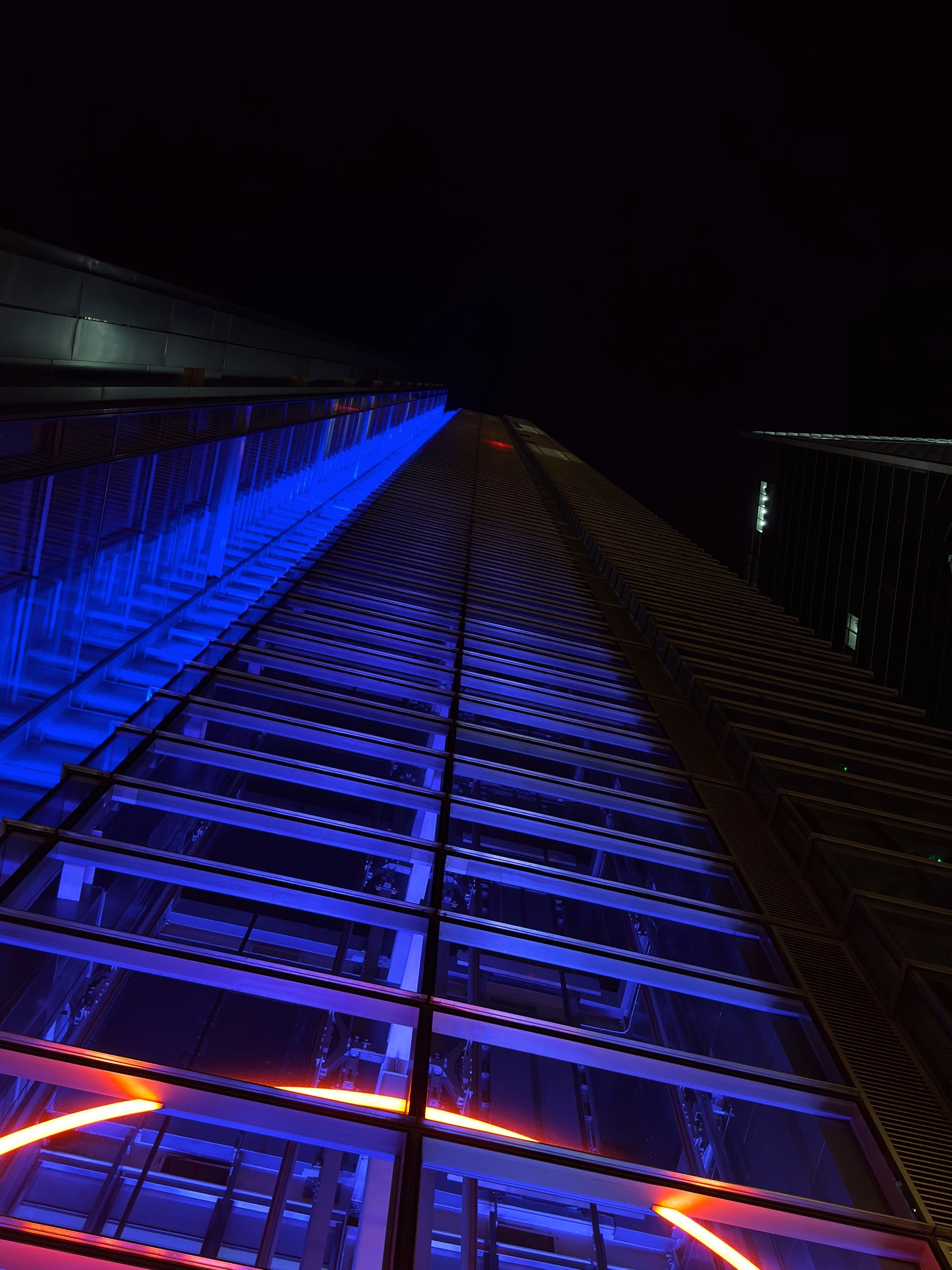 Upward-looking view along a modern glass-and-steel building, illuminated with blue accent lighting that emphasizes strong lines and geometric repetition against a black night sky.