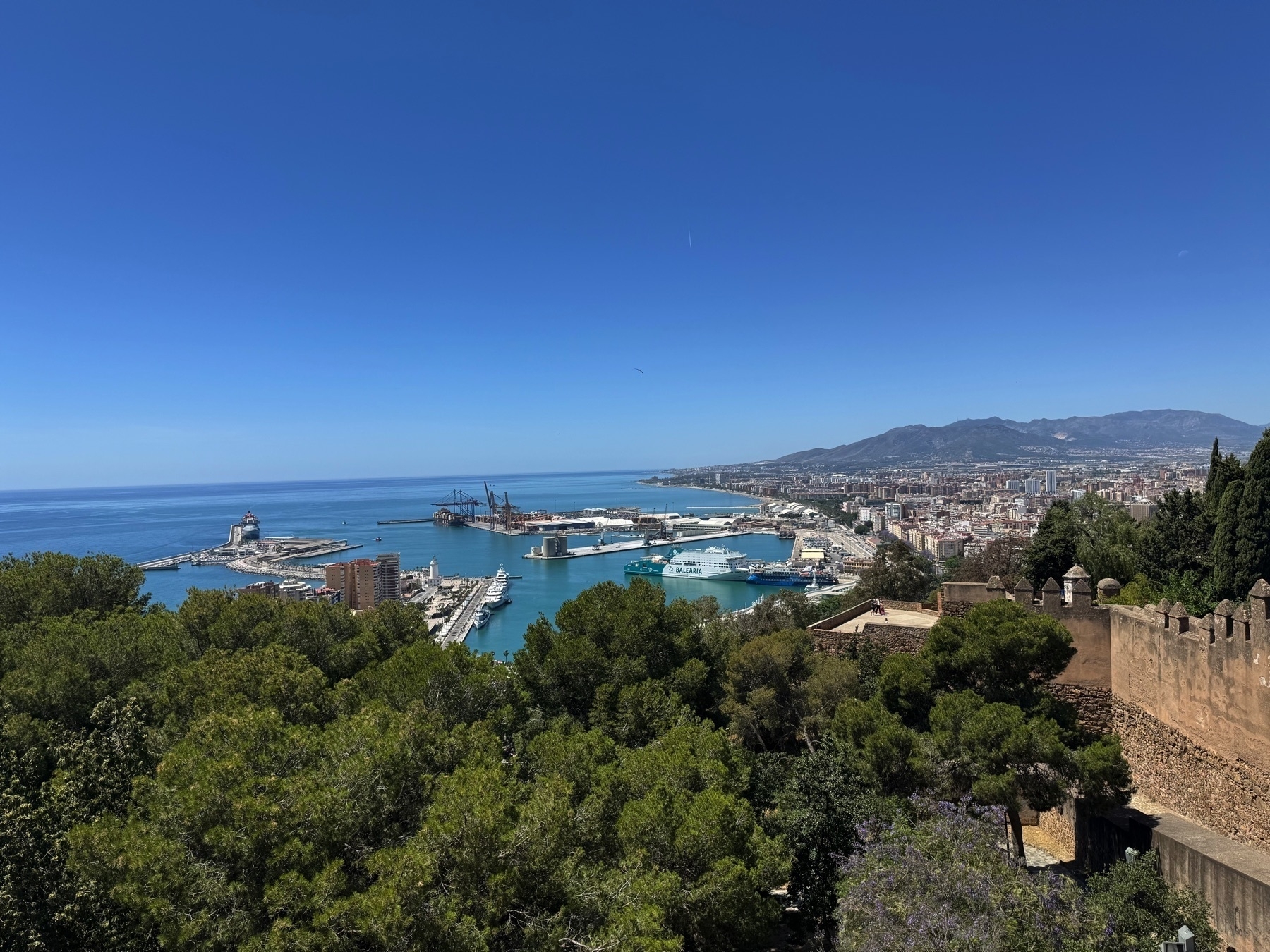 A sweeping view of Málaga’s harbor with ships docked along the waterfront and a tree-lined hillside leading down to the city. The azure sea meets the skyline under a pristine blue sky, framed by fortress walls.