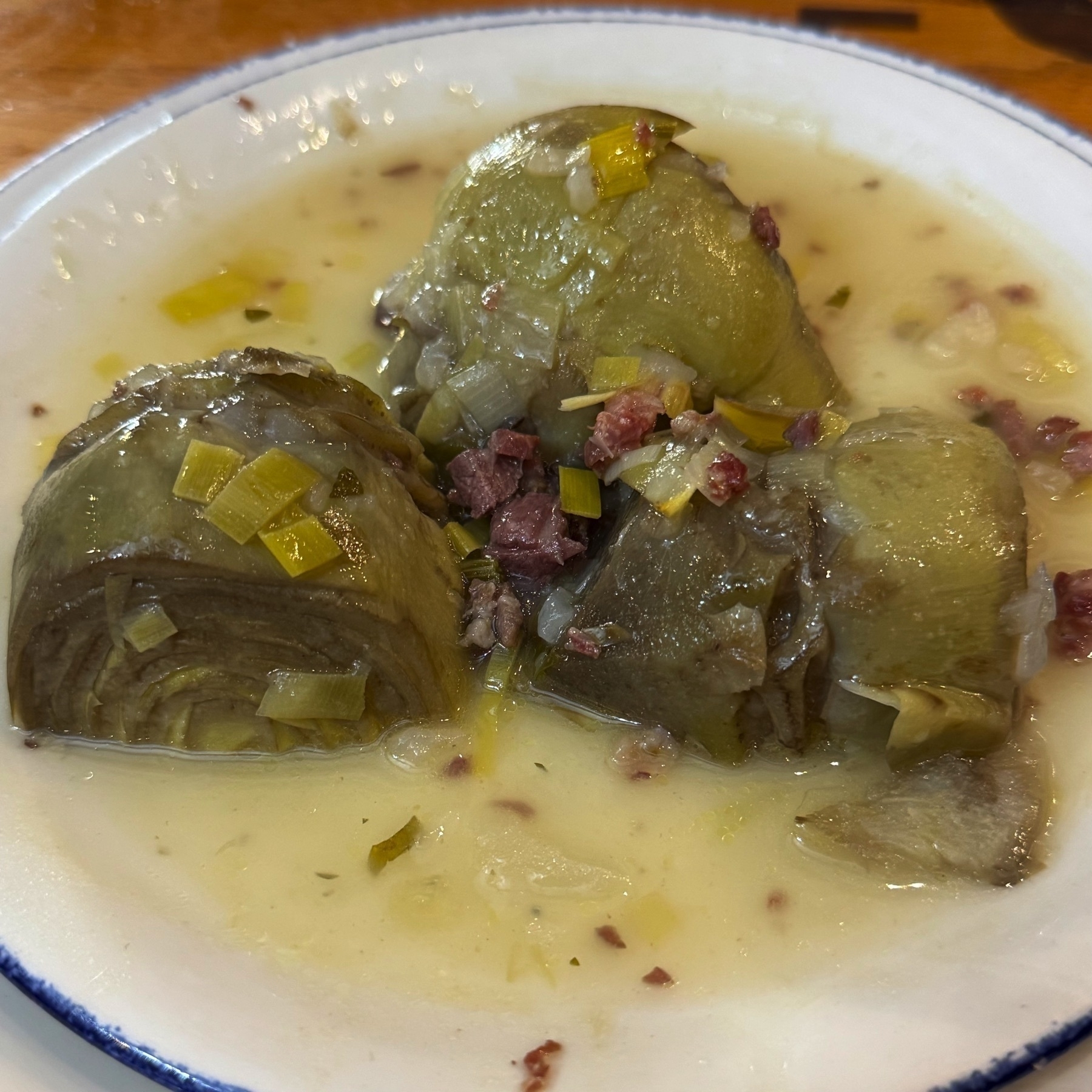 Halved artichoke hearts swimming in a light, creamy broth speckled with chopped ham and diced leeks. The broth clings to the vegetables, and the tender leaves show subtle layering, suggesting a soft bite and deep flavor.