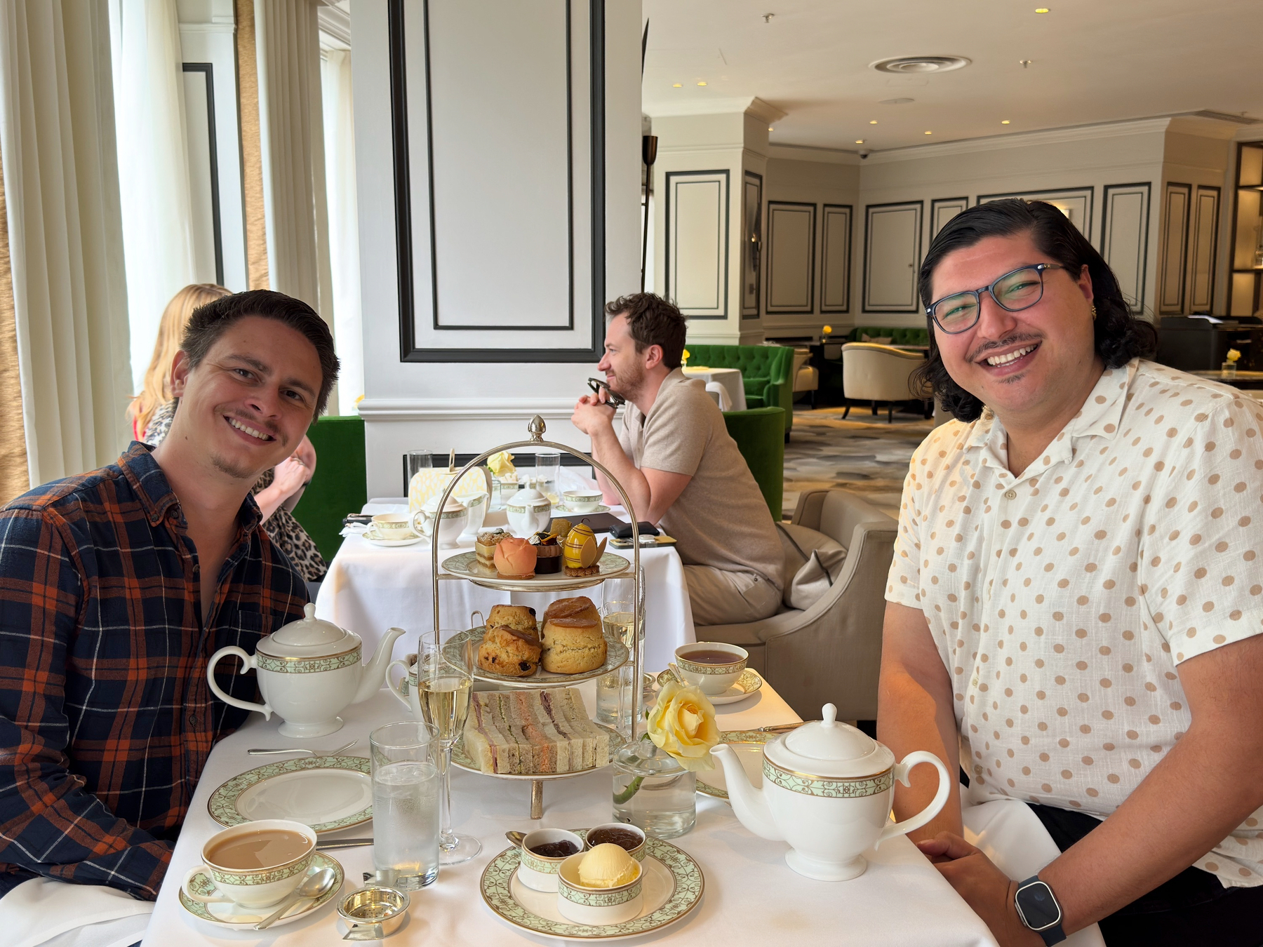Hugo and I seated around a white linen table set for high tea, with fine china, polished silverware, and a tiered stand of sandwiches, scones, and pastries. Soft daylight, green velvet chairs, and classic paneling give the room an elegant, relaxed feel.