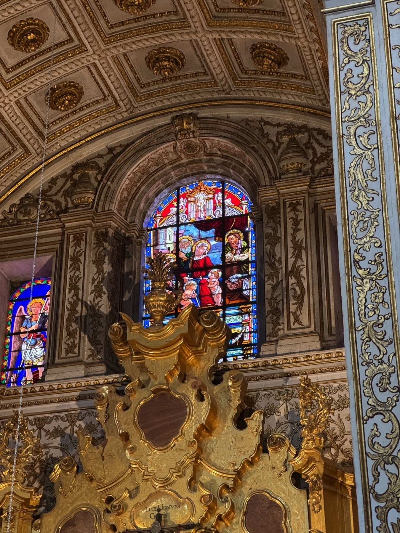 Close-up view of a stained-glass window set into an ornately decorated stone wall. The vibrant panel depicts religious figures in richly colored robes, their expressions solemn and reverent. Below the window, a gilded architectural element with curving forms and rounded plaques adds depth and texture to the composition.